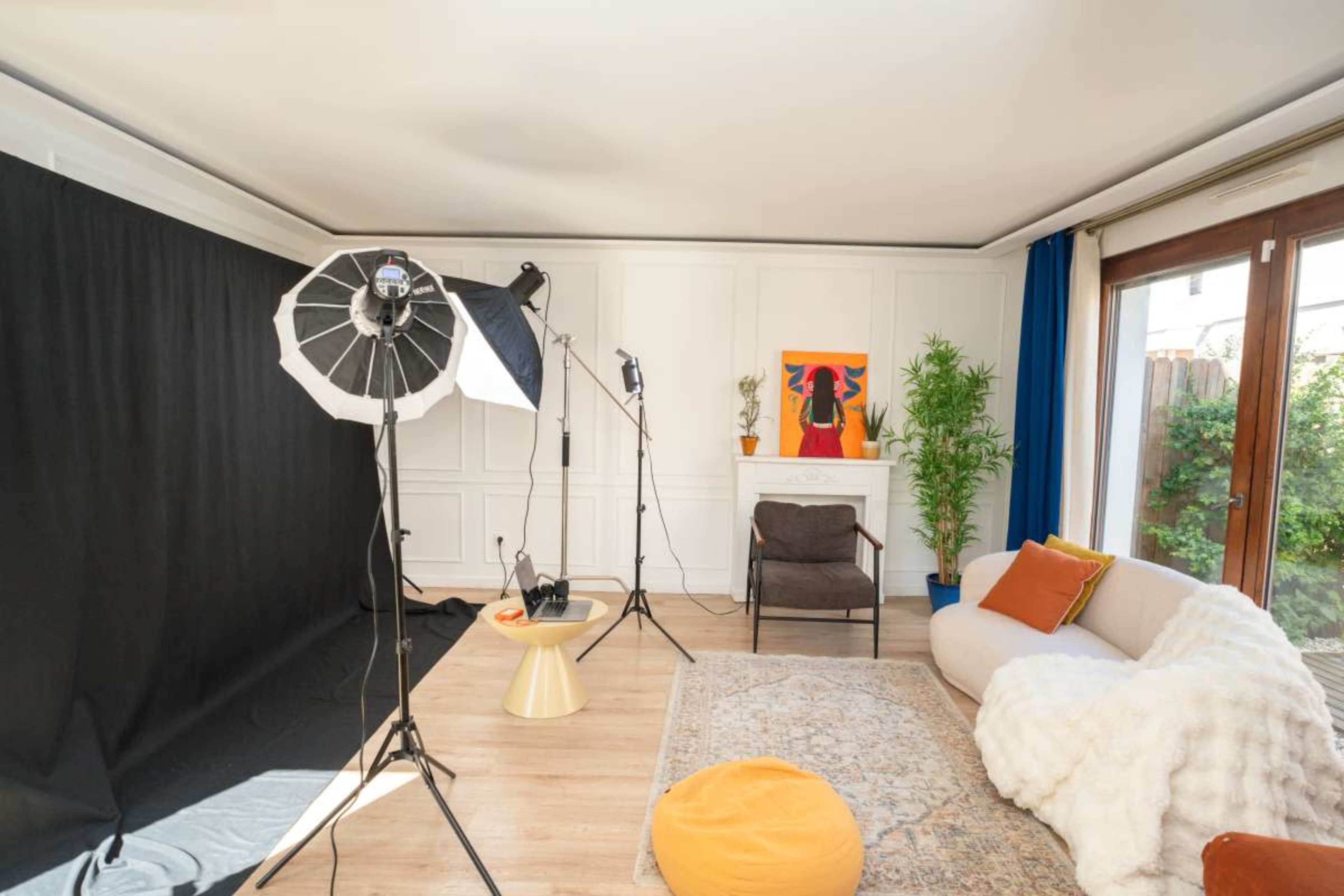 The image shows a well-lit photo studio setup featuring a black backdrop, lighting equipment, a cozy sofa, and decorative plants.