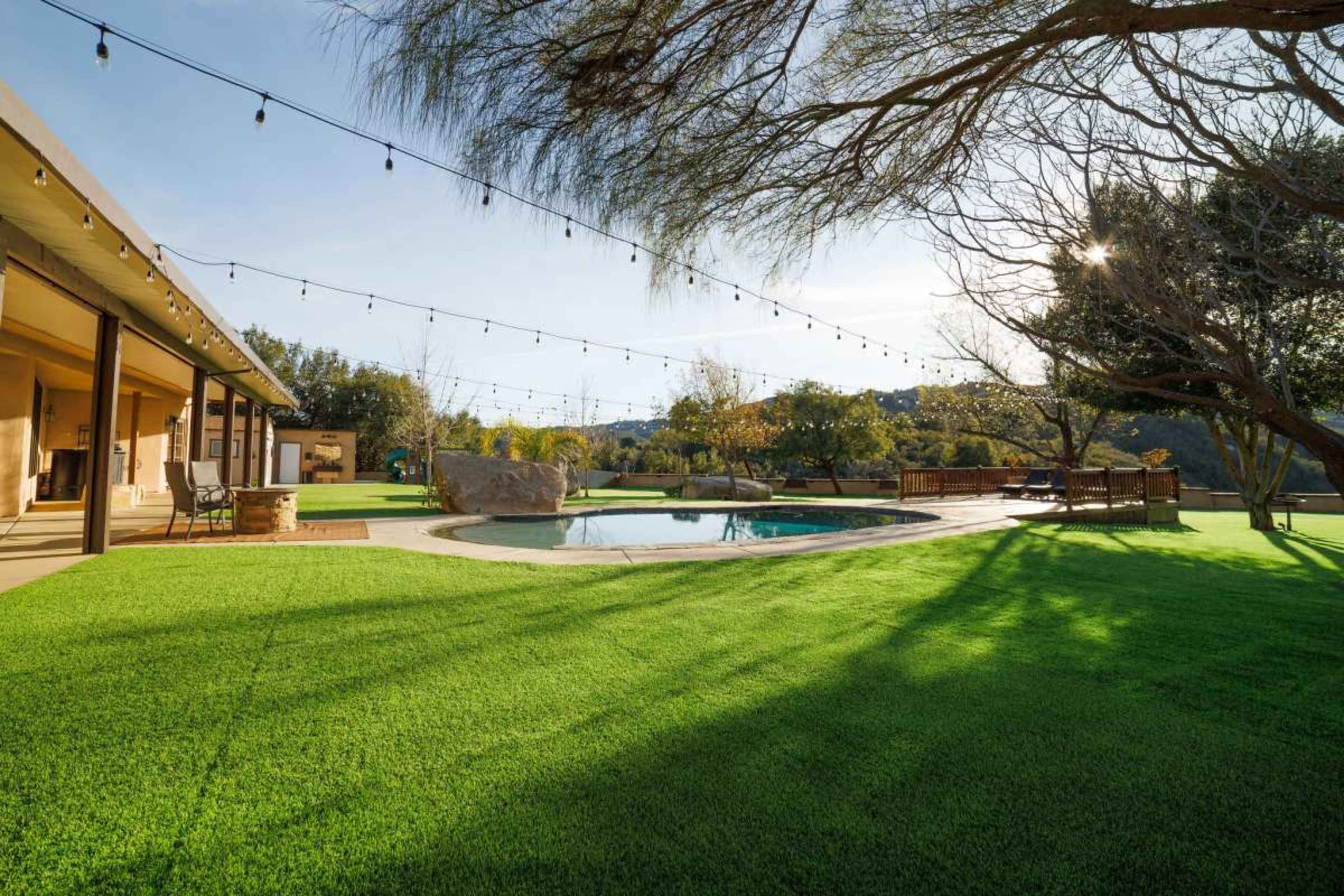 The image shows a backyard with a swimming pool, surrounded by manicured artificial grass, a large rock, and several trees under string lights in a sunny setting.