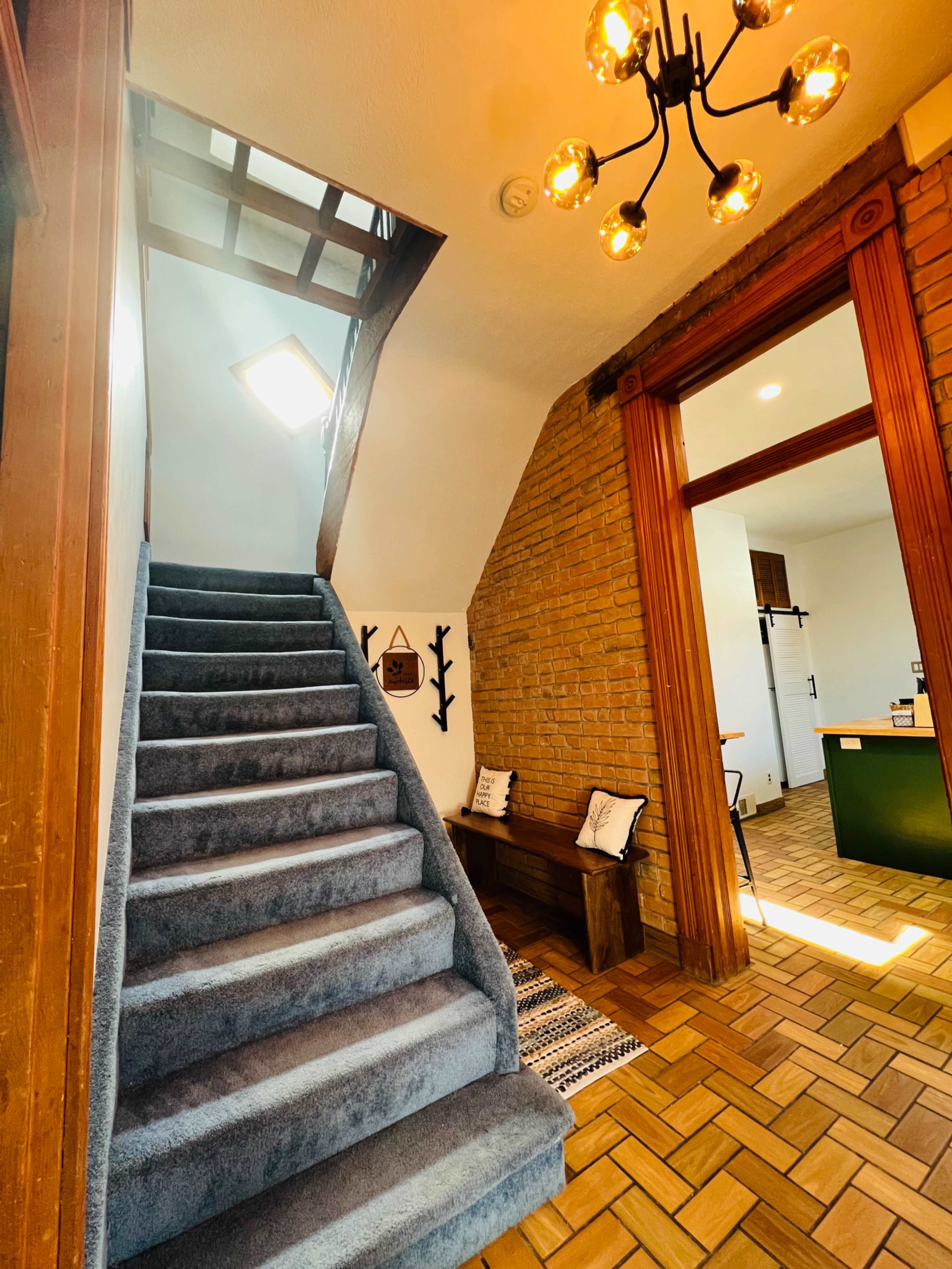 A staircase with blue carpet leading to a second floor, flanked by a brick wall on one side and a wooden door on the other.