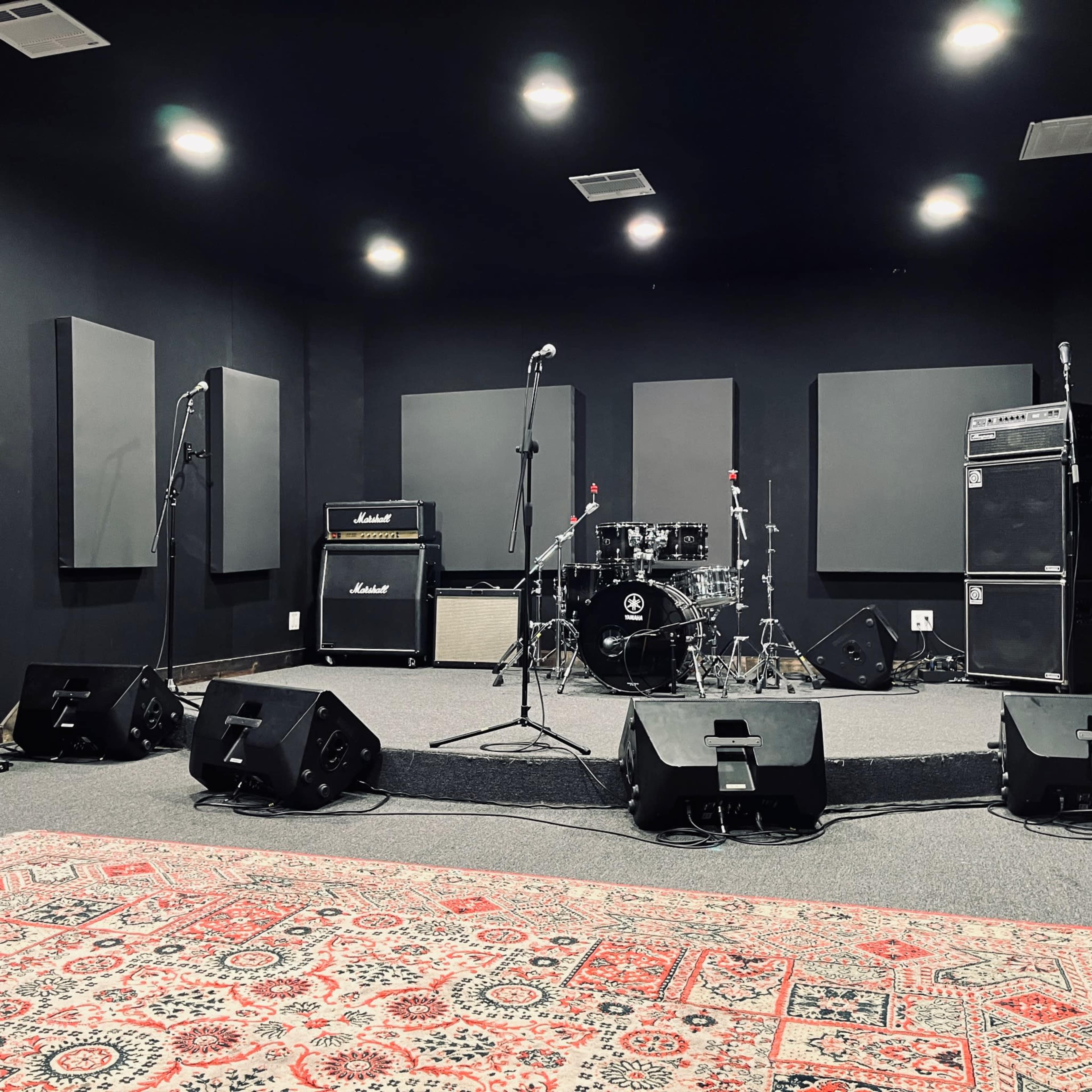 The image shows a music rehearsal space with a drum kit, amplifiers, microphones, and sound equipment set up on a raised platform.