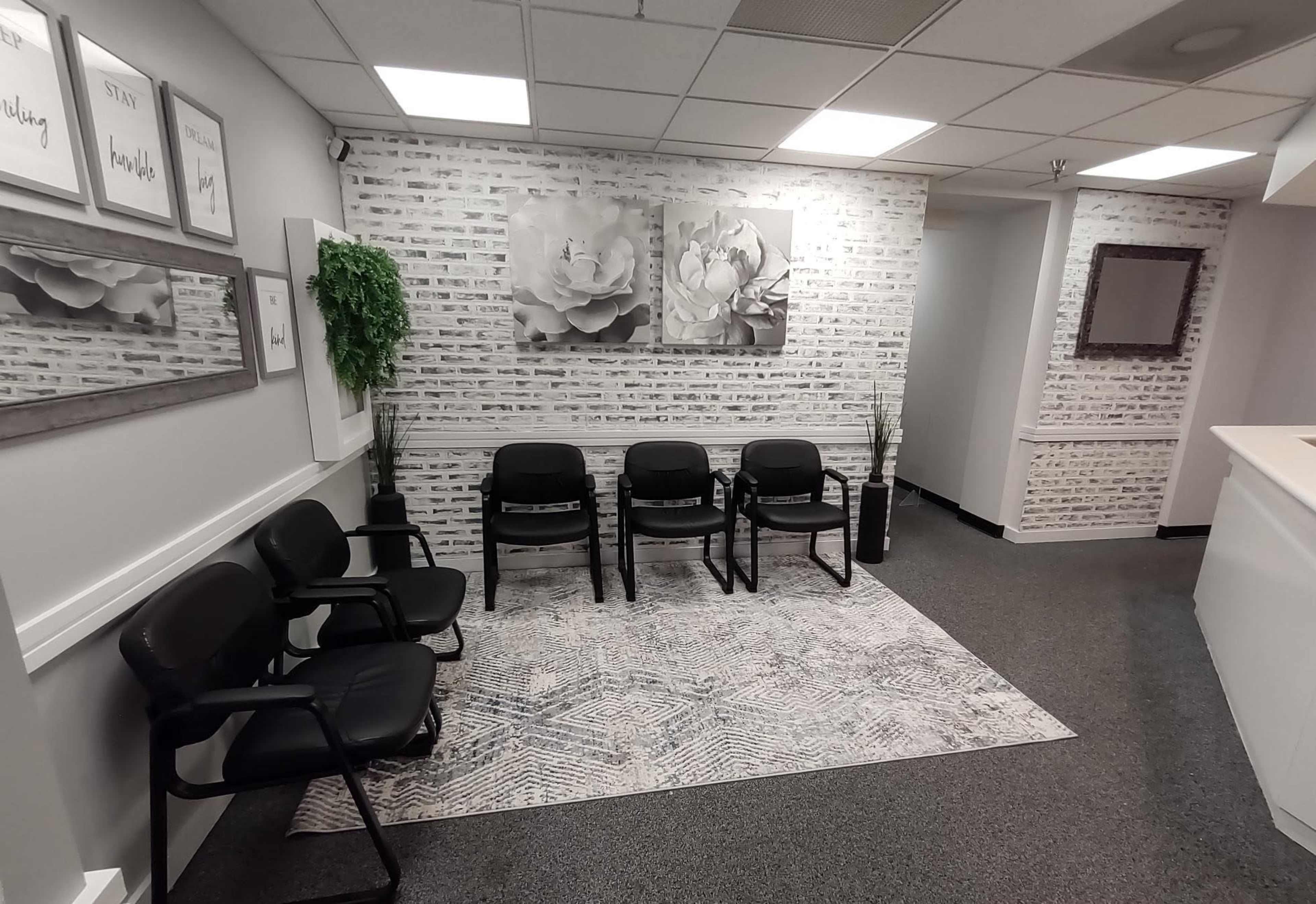 The image shows a waiting area with four black chairs, decorative wall art featuring flowers, and a potted plant.