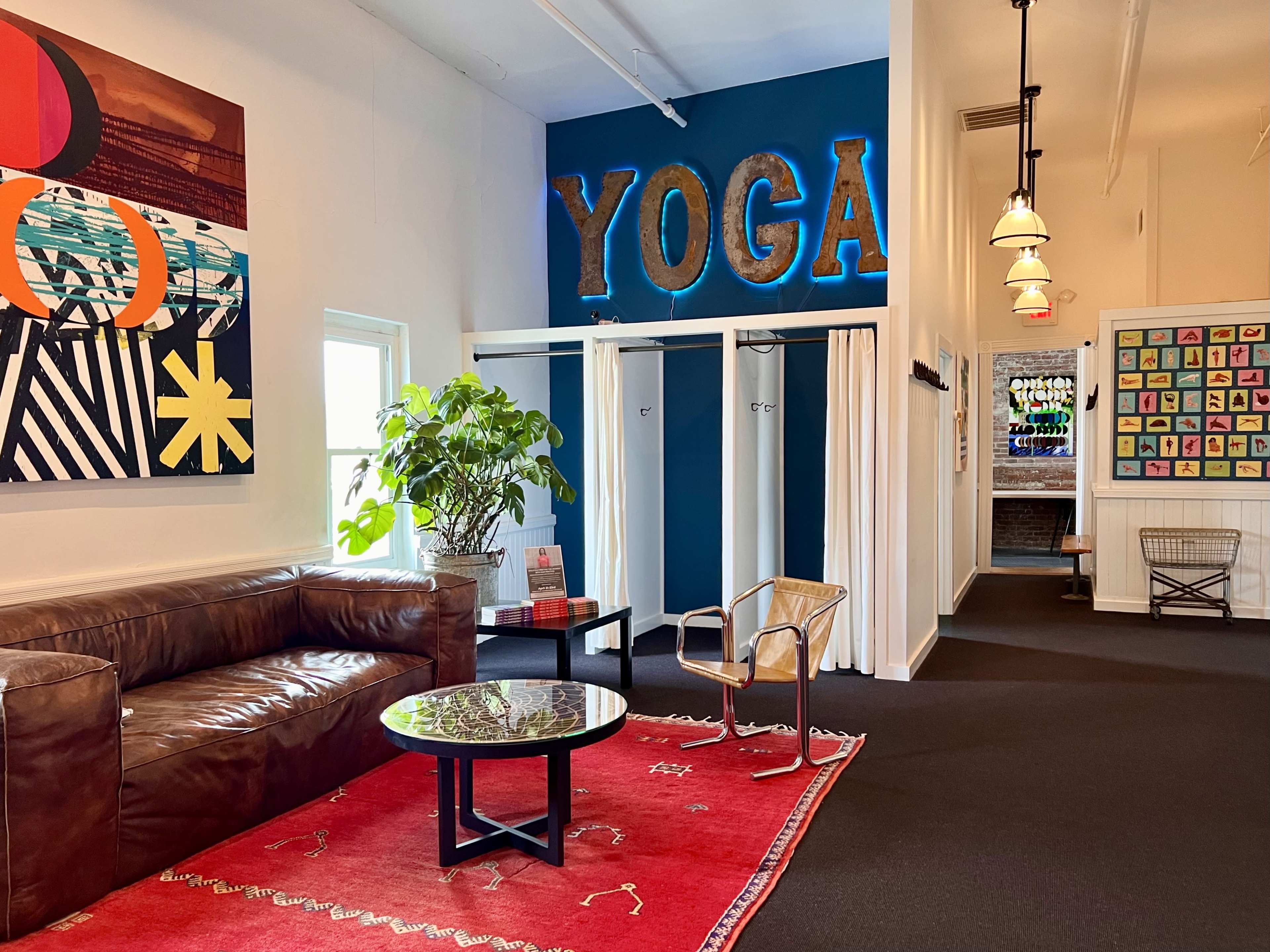 A spacious interior of a yoga studio, featuring a cozy seating area with a brown leather sofa and a round table, alongside a vibrant "YOGA" sign on a blue wall and changing rooms with white curtains.