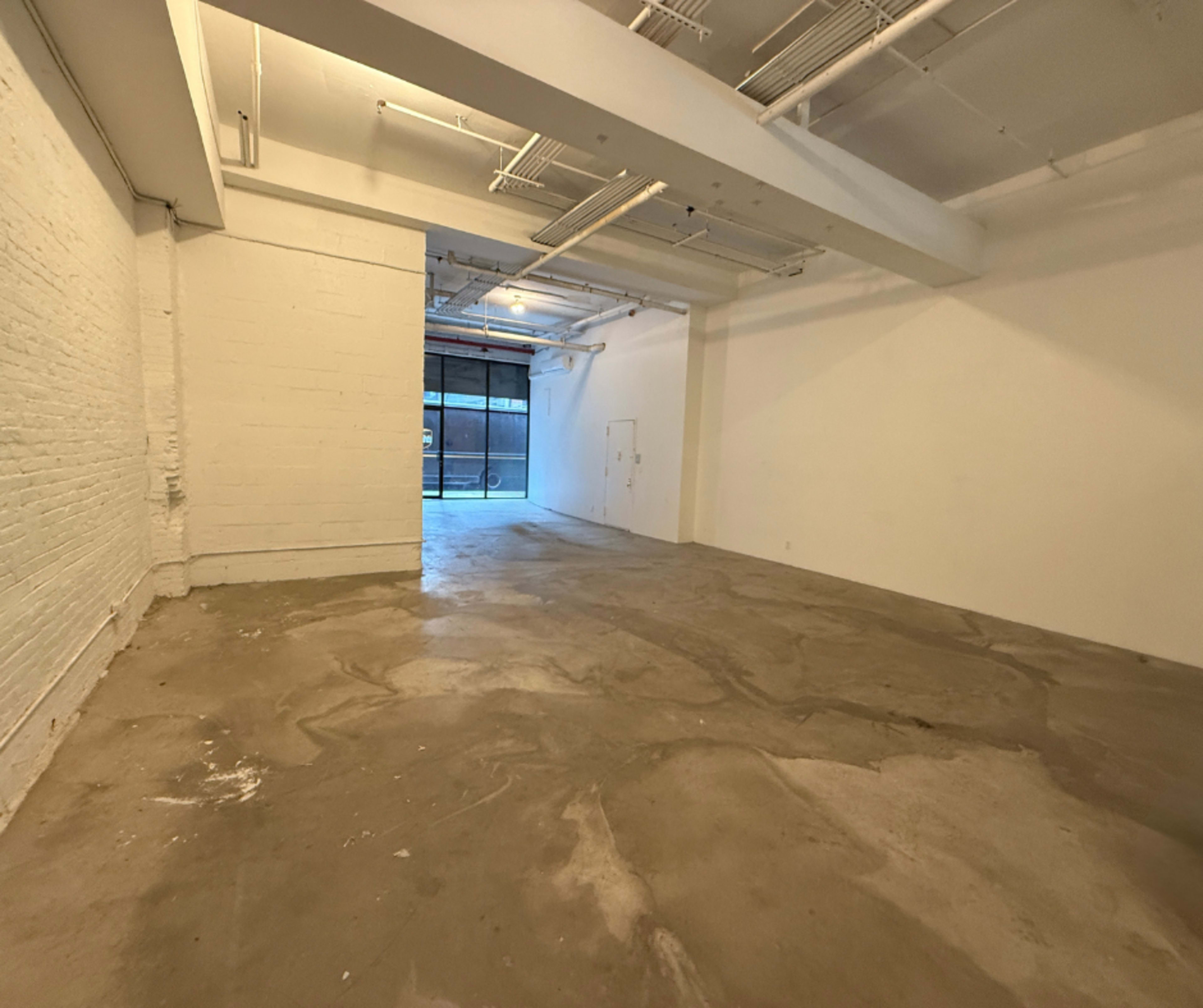 The image shows a spacious, empty interior with concrete floors and white walls, leading to a large opening that provides natural light from outside.