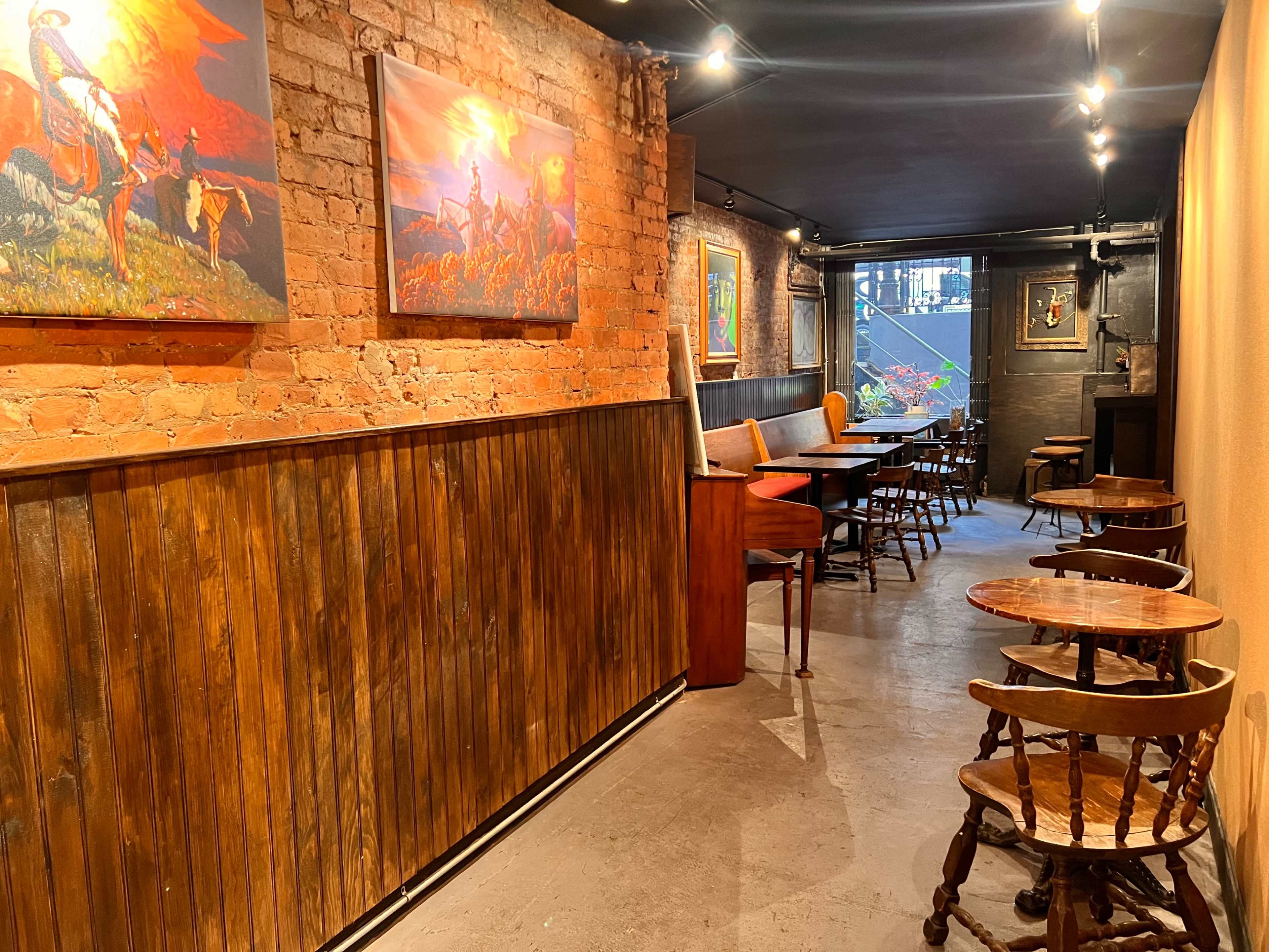 The image shows a narrow, well-lit corridor in a rustic restaurant with wooden walls, featuring art on the brick wall and seating areas along the sides.