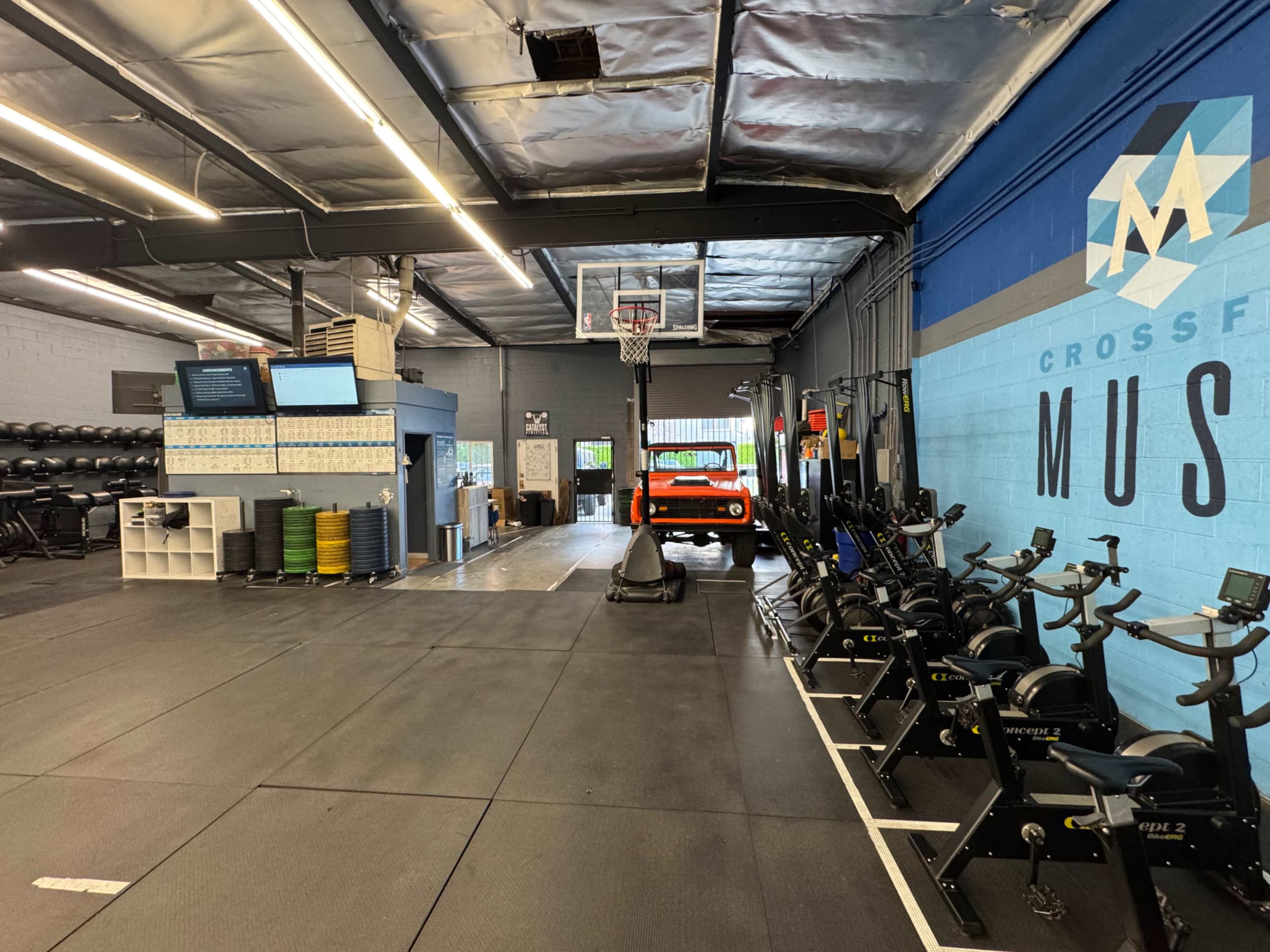 The image shows a spacious gym interior with exercise equipment, a basketball hoop, and a red vehicle parked in the background.