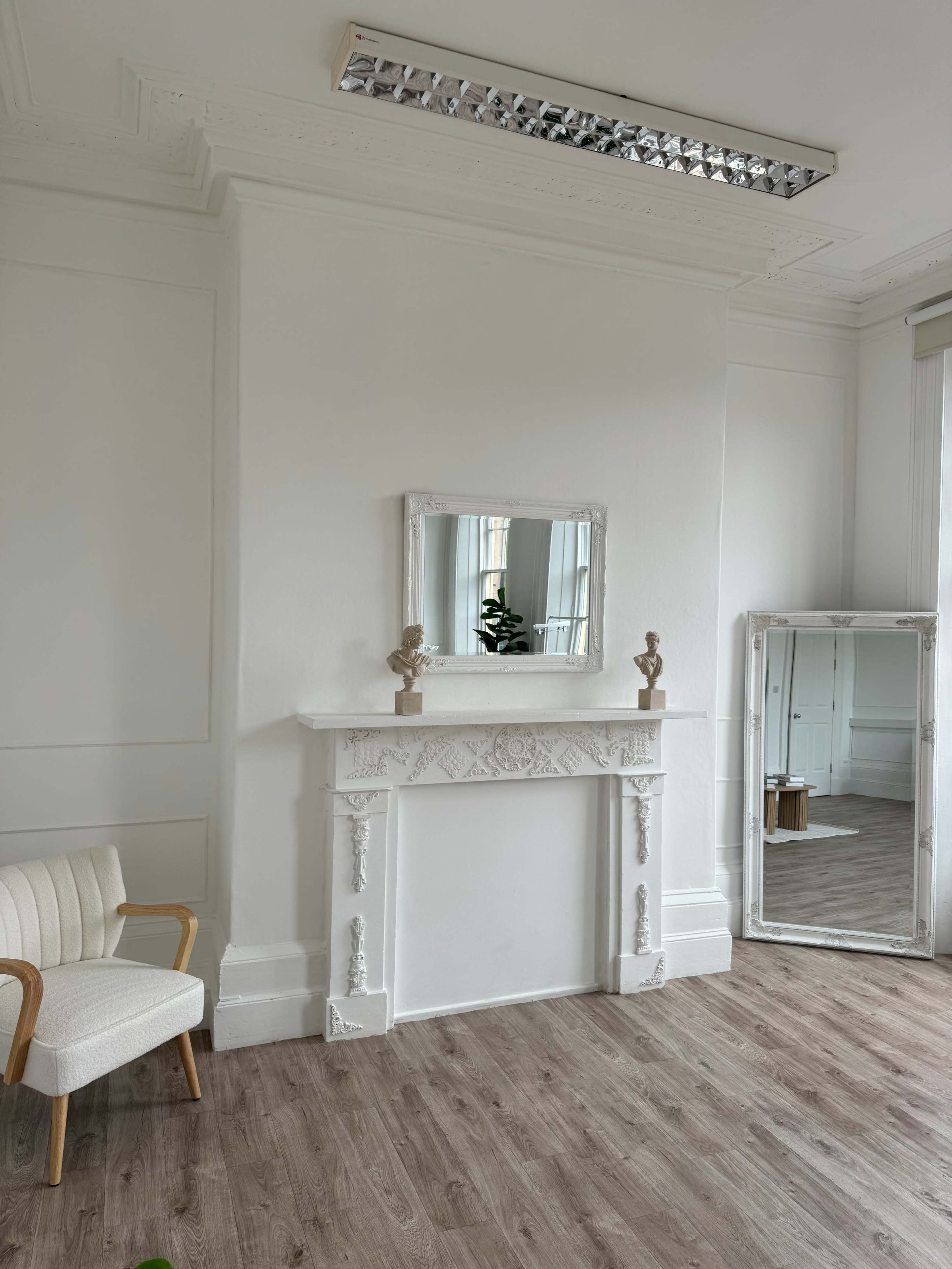 The image shows a bright room with white walls, a vintage fireplace with ornate detailing, a mirror above it, a modern chair to the left, and a large mirror leaning against the right wall.