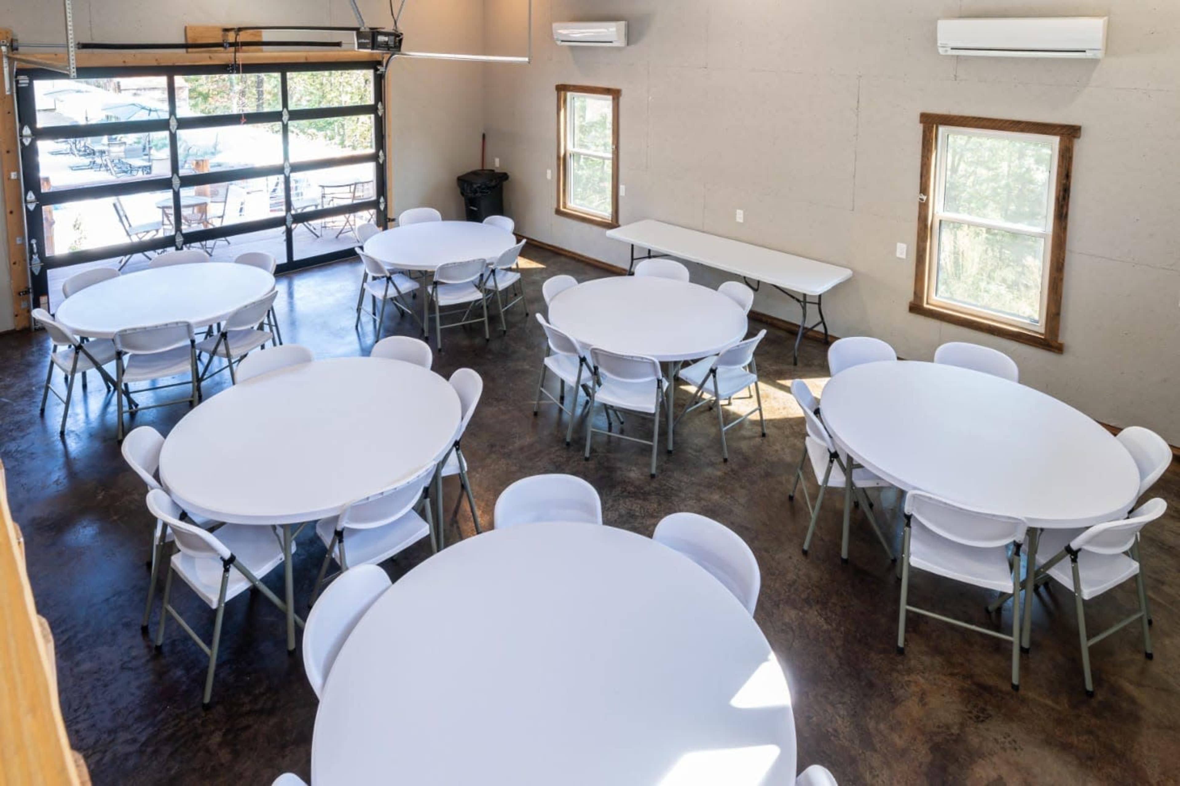 A spacious room with multiple round tables and chairs arranged neatly, along with large windows allowing natural light.
