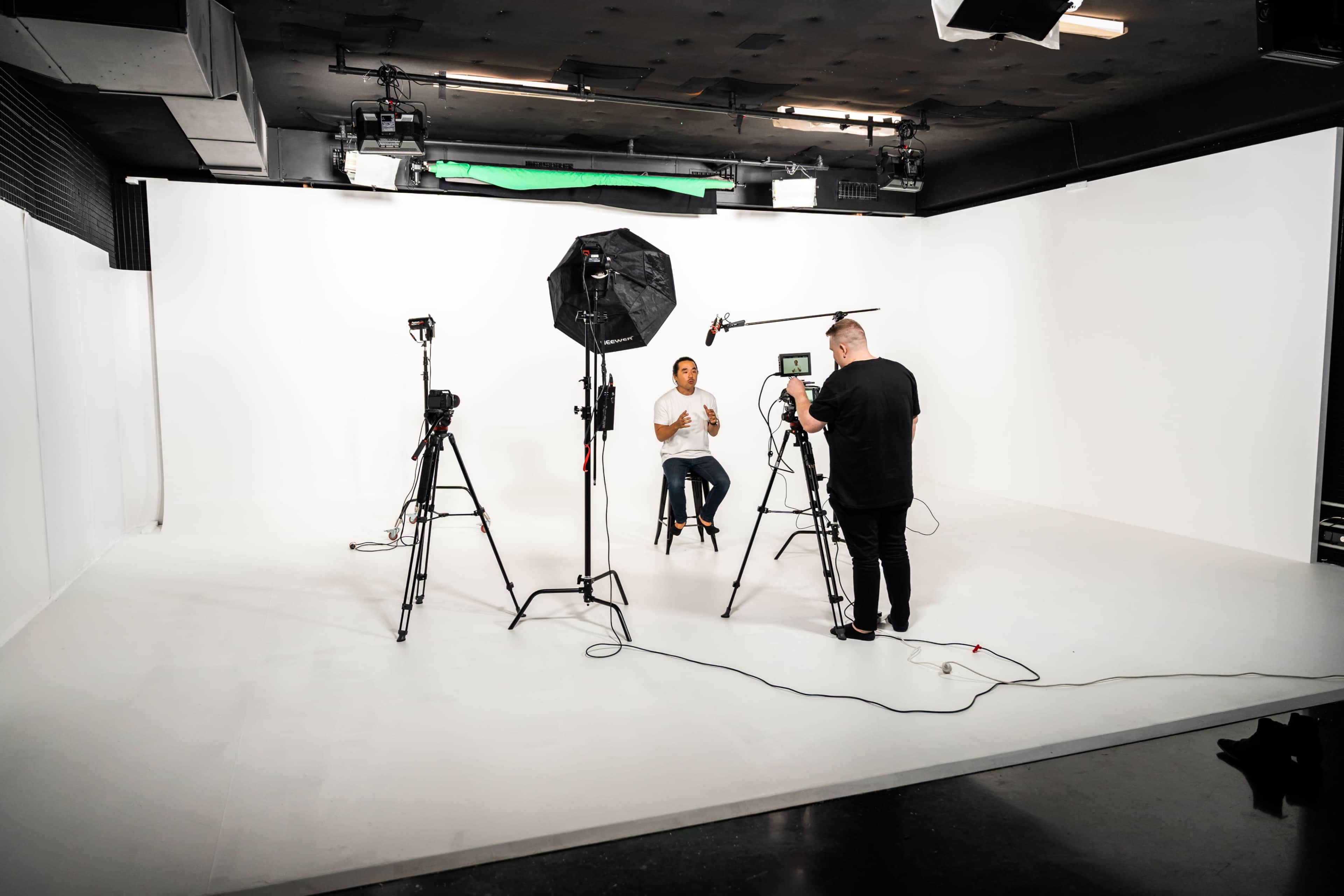 A person seated on a stool is being interviewed by another person in a studio with a white background, surrounded by cameras and lighting equipment.