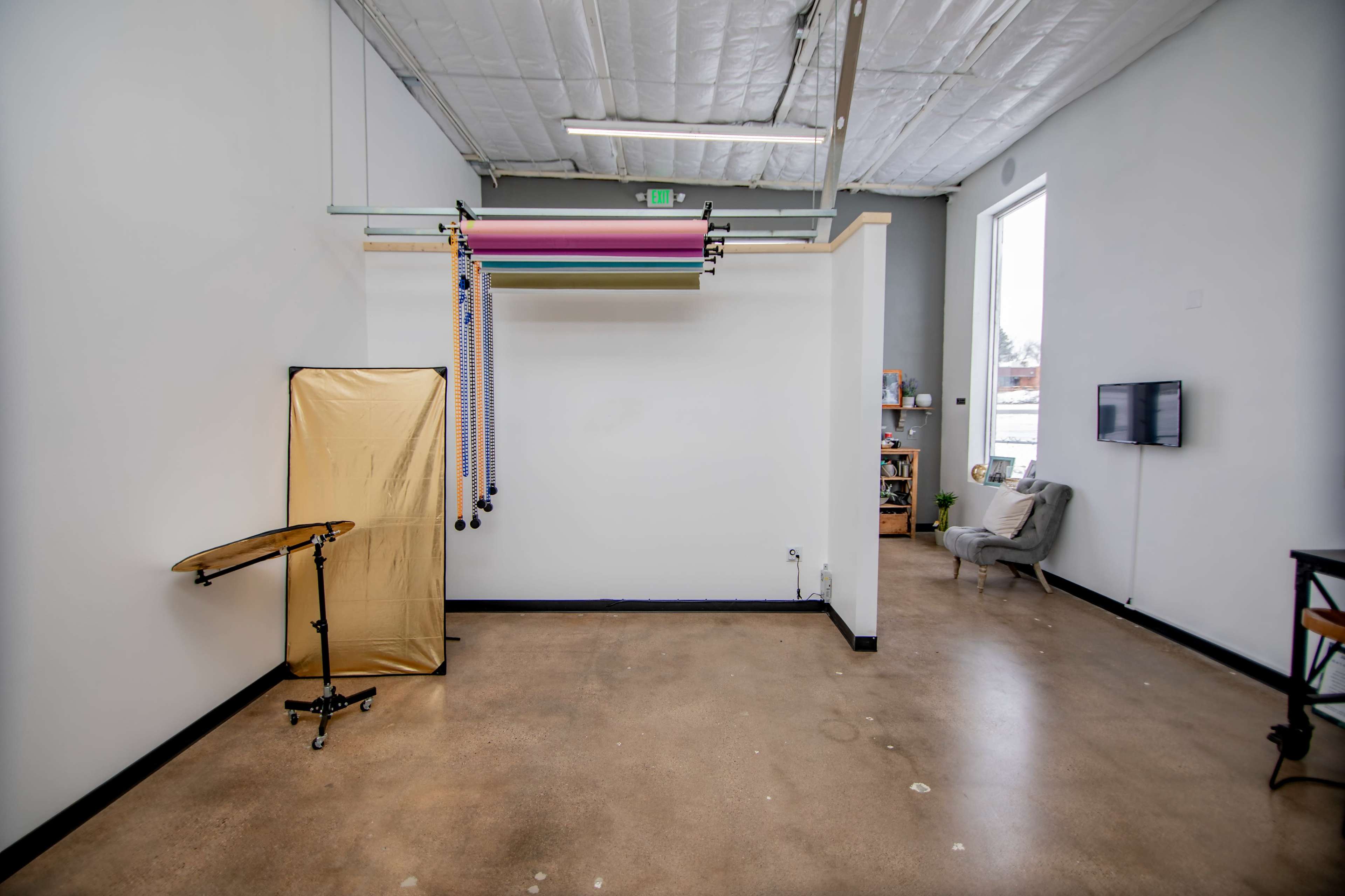 The image shows an empty studio space with a gold backdrop stand, hanging colorful backdrops, and minimal furnishings.