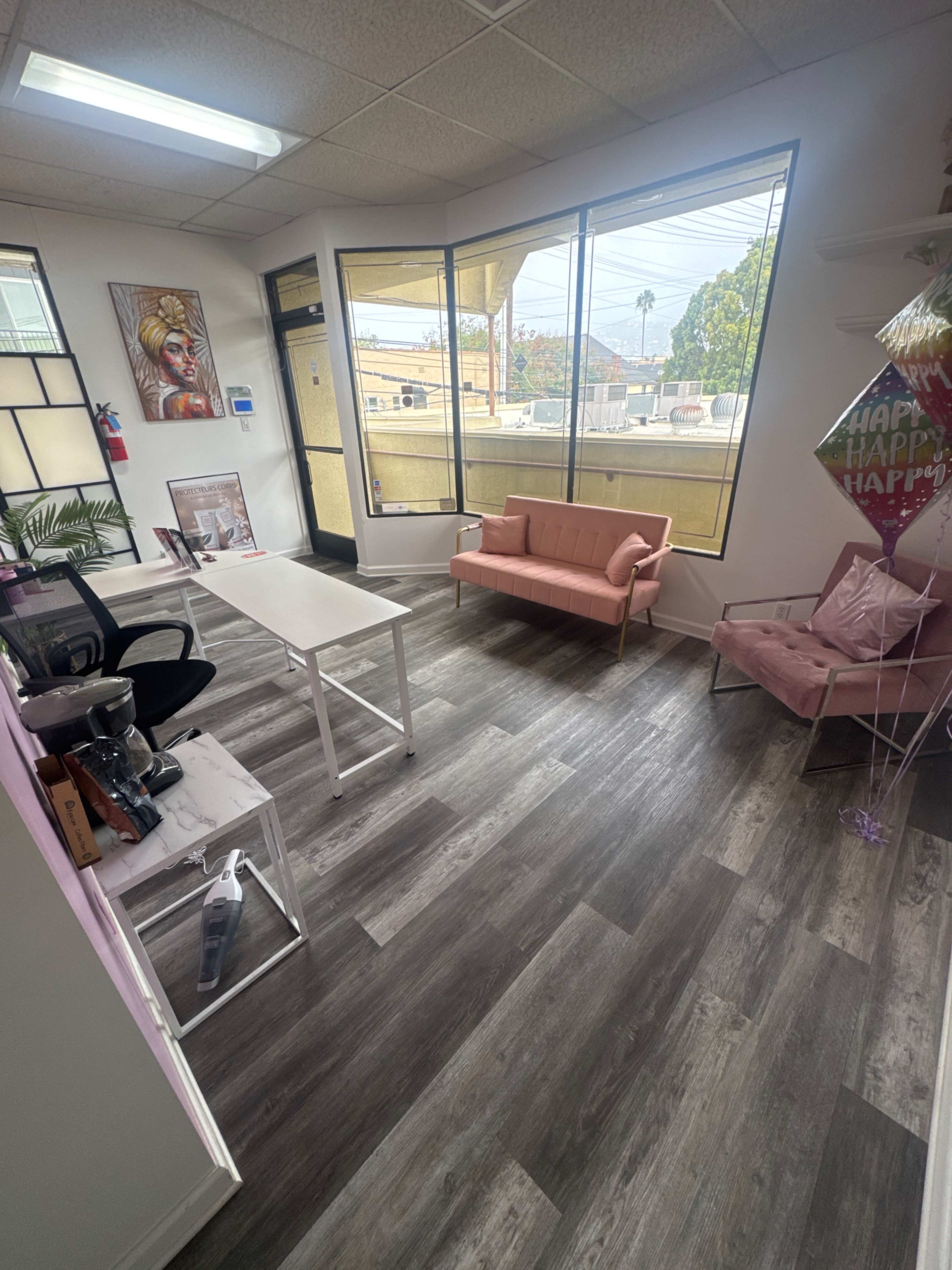 The image shows a modern office interior with large windows, a pink sofa, and a white table surrounded by light wood flooring.