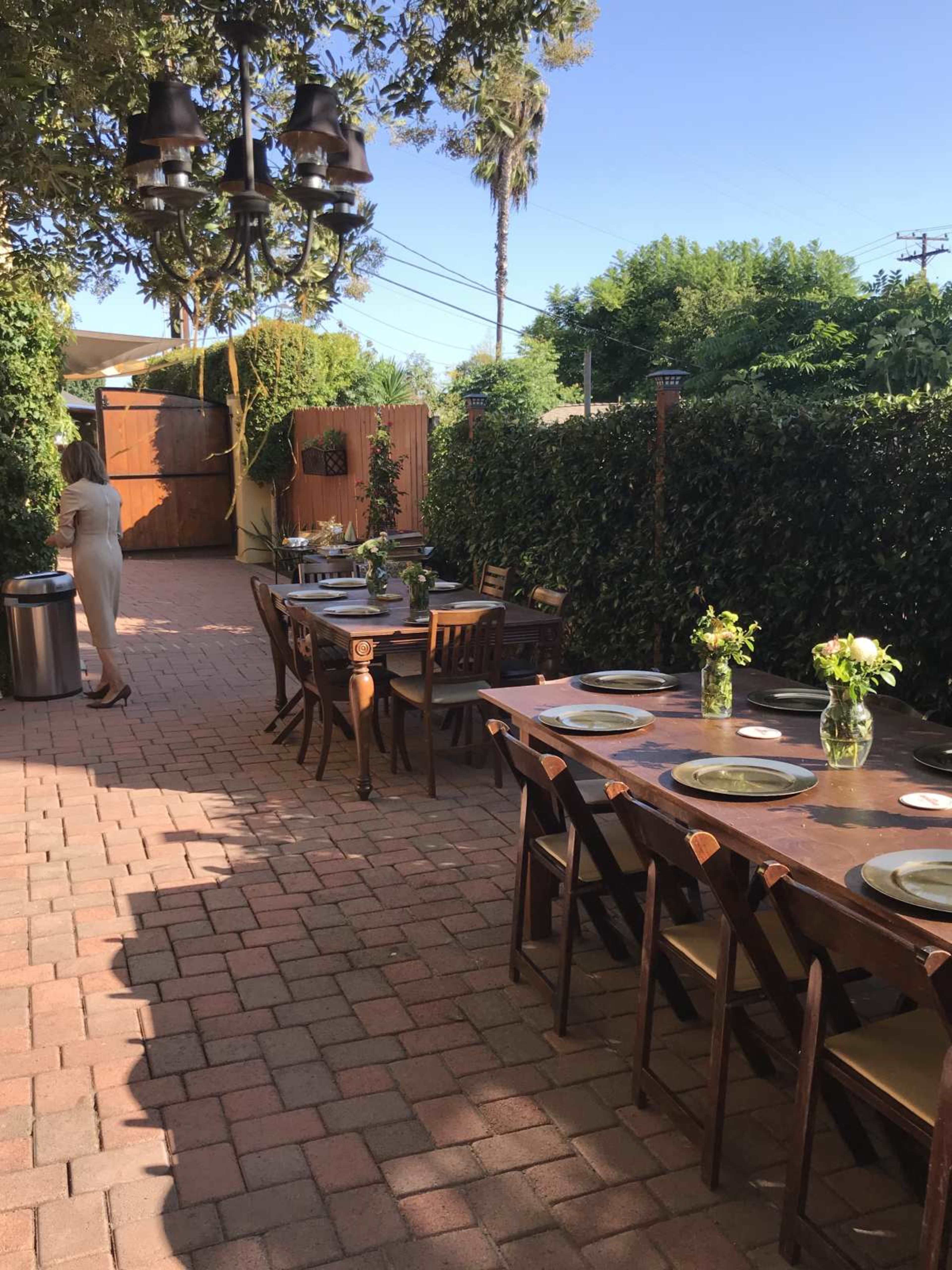 A long outdoor dining table set with flower centerpieces is arranged in a patio bordered by hedges, with a woman standing nearby.