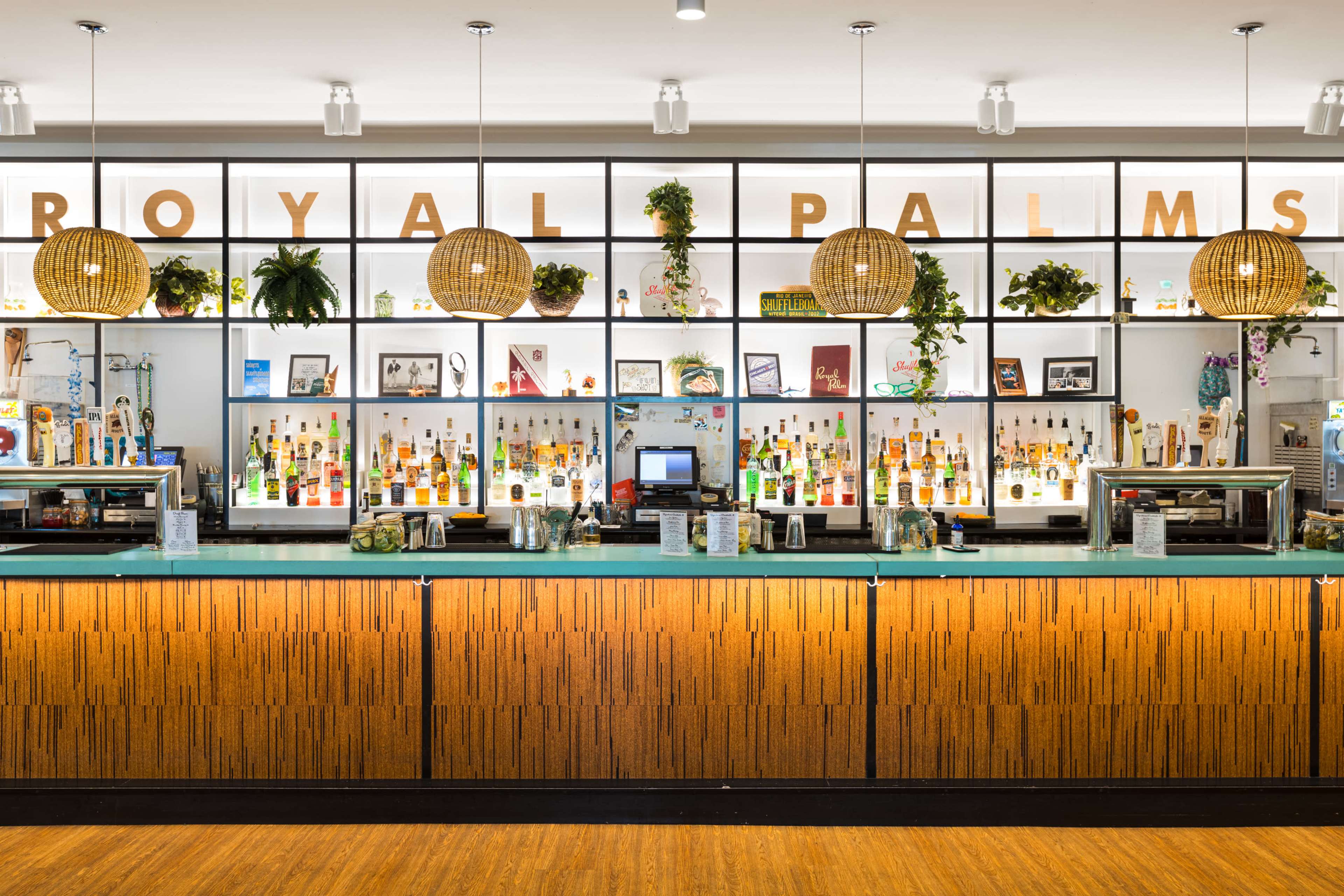 The image shows a bar area with a well-organized display of various bottles, decorative plants, and a large sign reading "Royal Palms" above the counter.