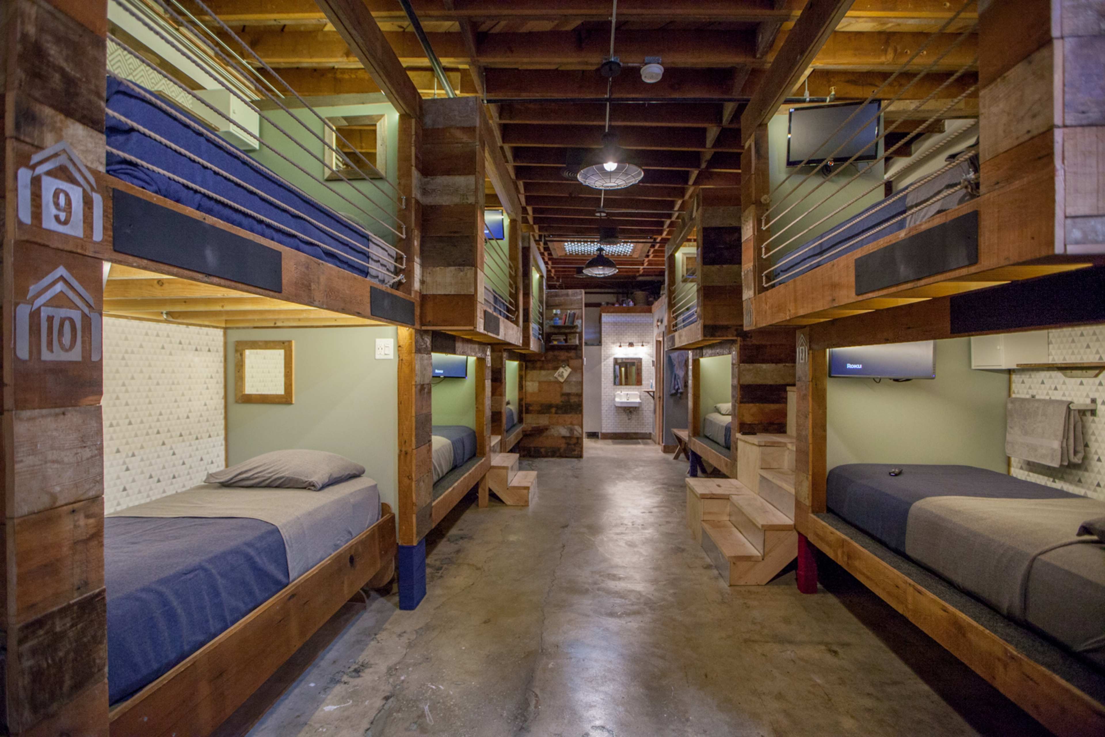 The image shows a modern shared dormitory with wooden bunk beds arranged in two rows, each with a small TV and built-in storage, under a wooden ceiling.