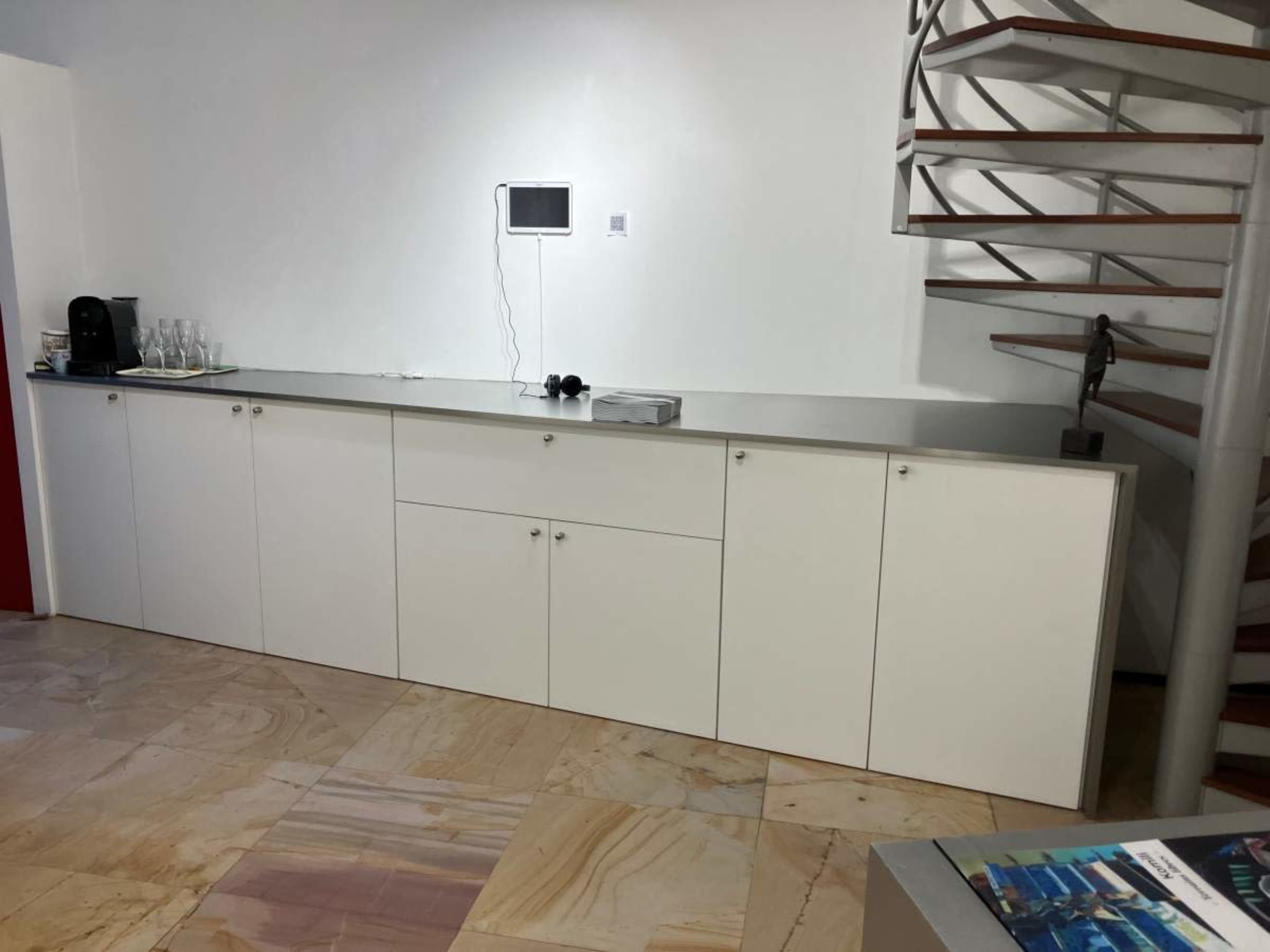 The image shows a minimalist kitchen area with a long white cabinetry countertop and a spiral staircase in the background.