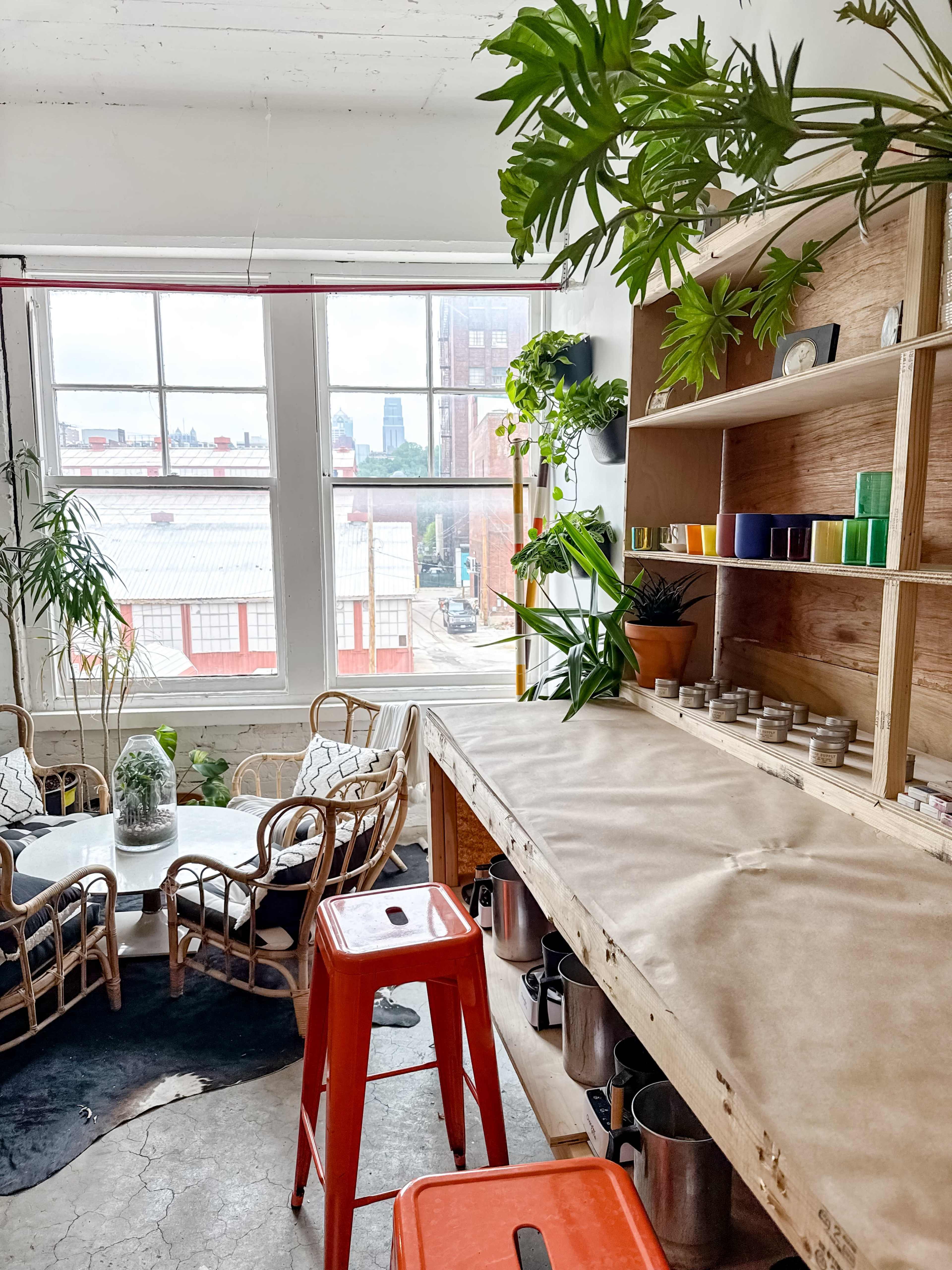 The image depicts a cozy interior space with a wooden shelf, orange stools, and a small table surrounded by plants, overlooking city buildings through large windows.