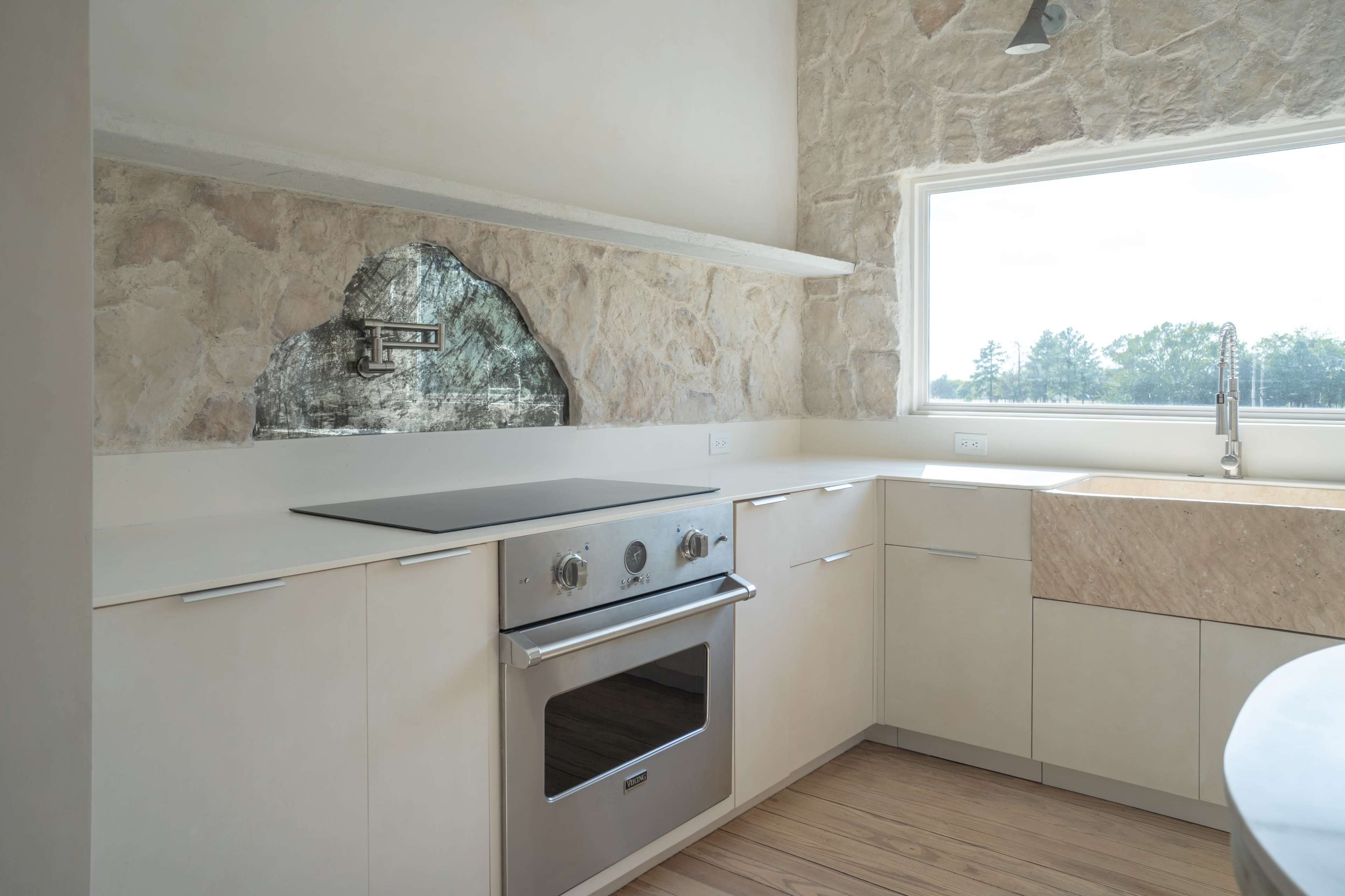 The kitchen features light-colored cabinets, a built-in oven, and a large window with a view, complemented by a stone wall and a unique stone shelf.
