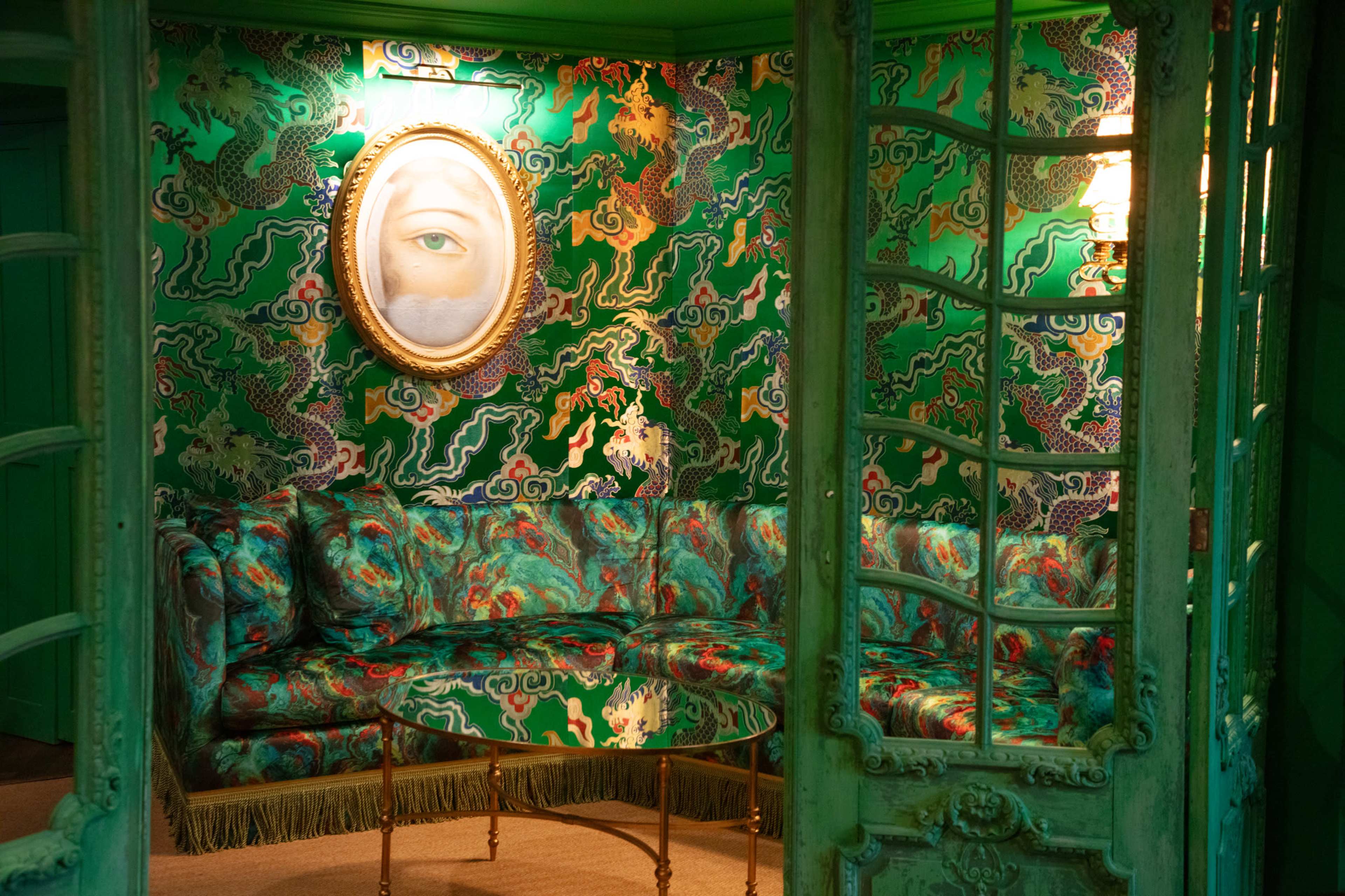 The image shows a vibrant green room featuring a patterned wallpaper, a circular mirror with an eye design, and a plush couch with intricate colors and designs.