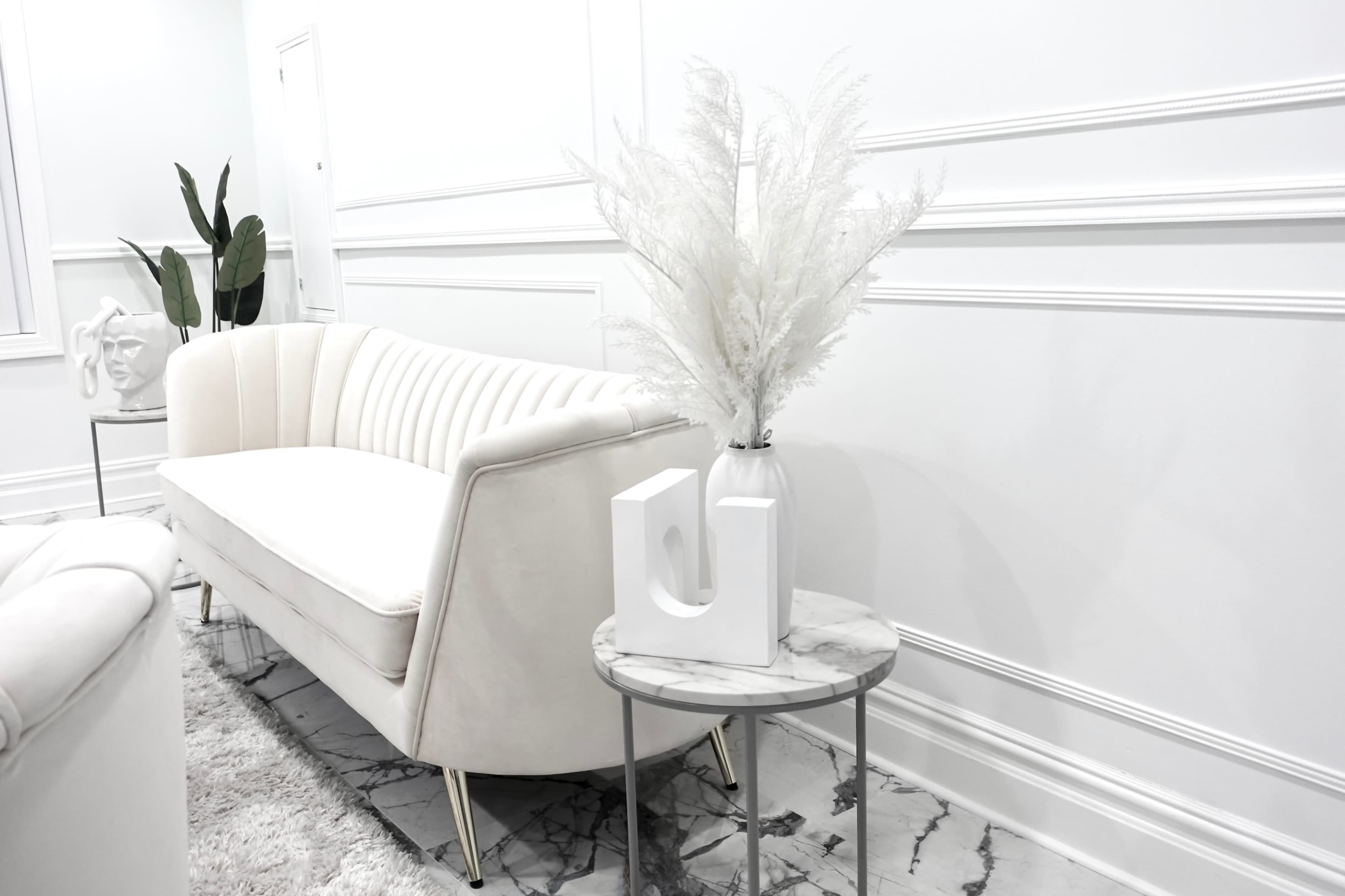 A white upholstered sofa is positioned next to a small marble-topped side table holding a minimalist white vase with decorative foliage.