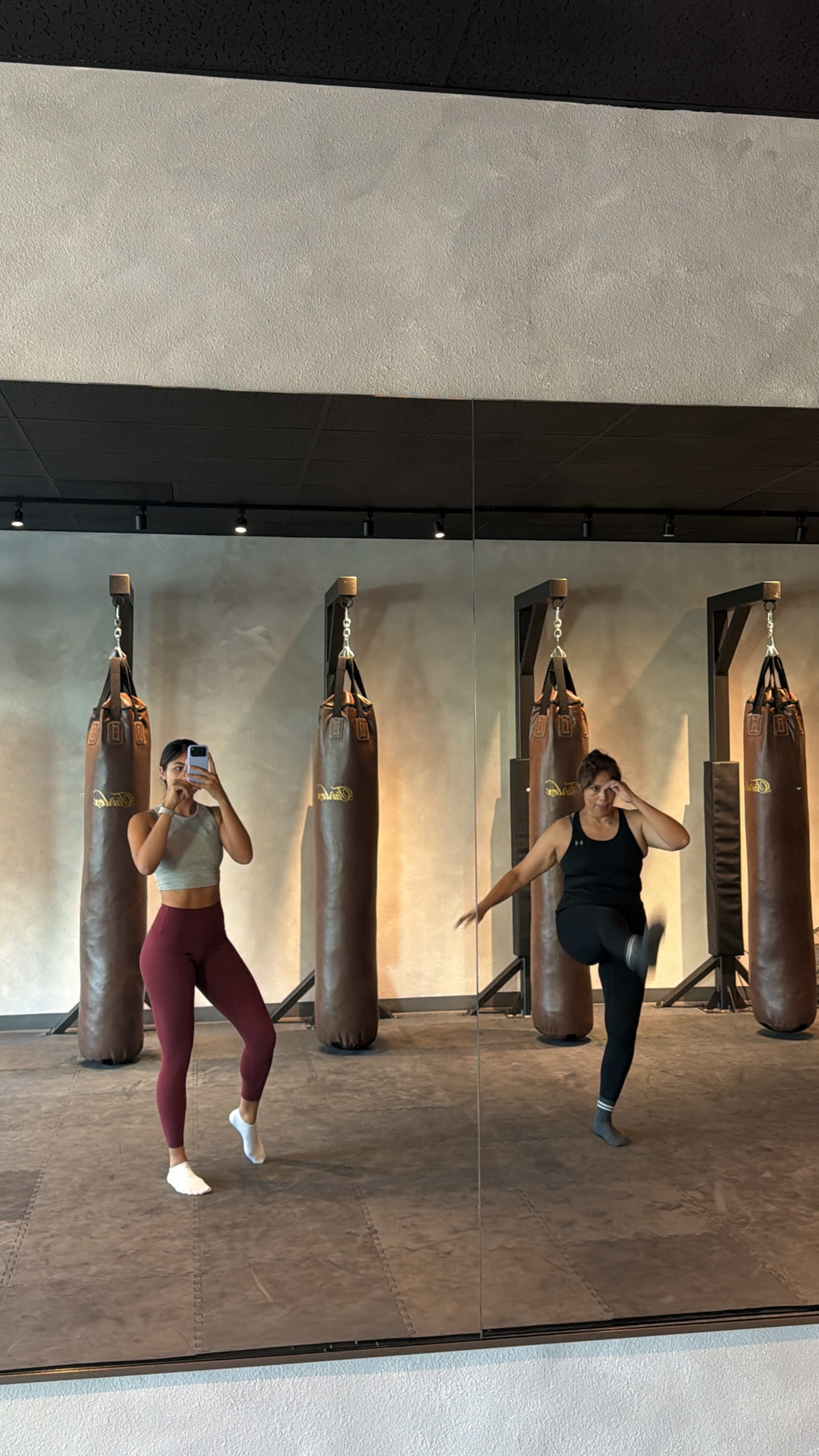 Two women are practicing workout routines in a gym, reflected in a mirror, with punching bags in the background.