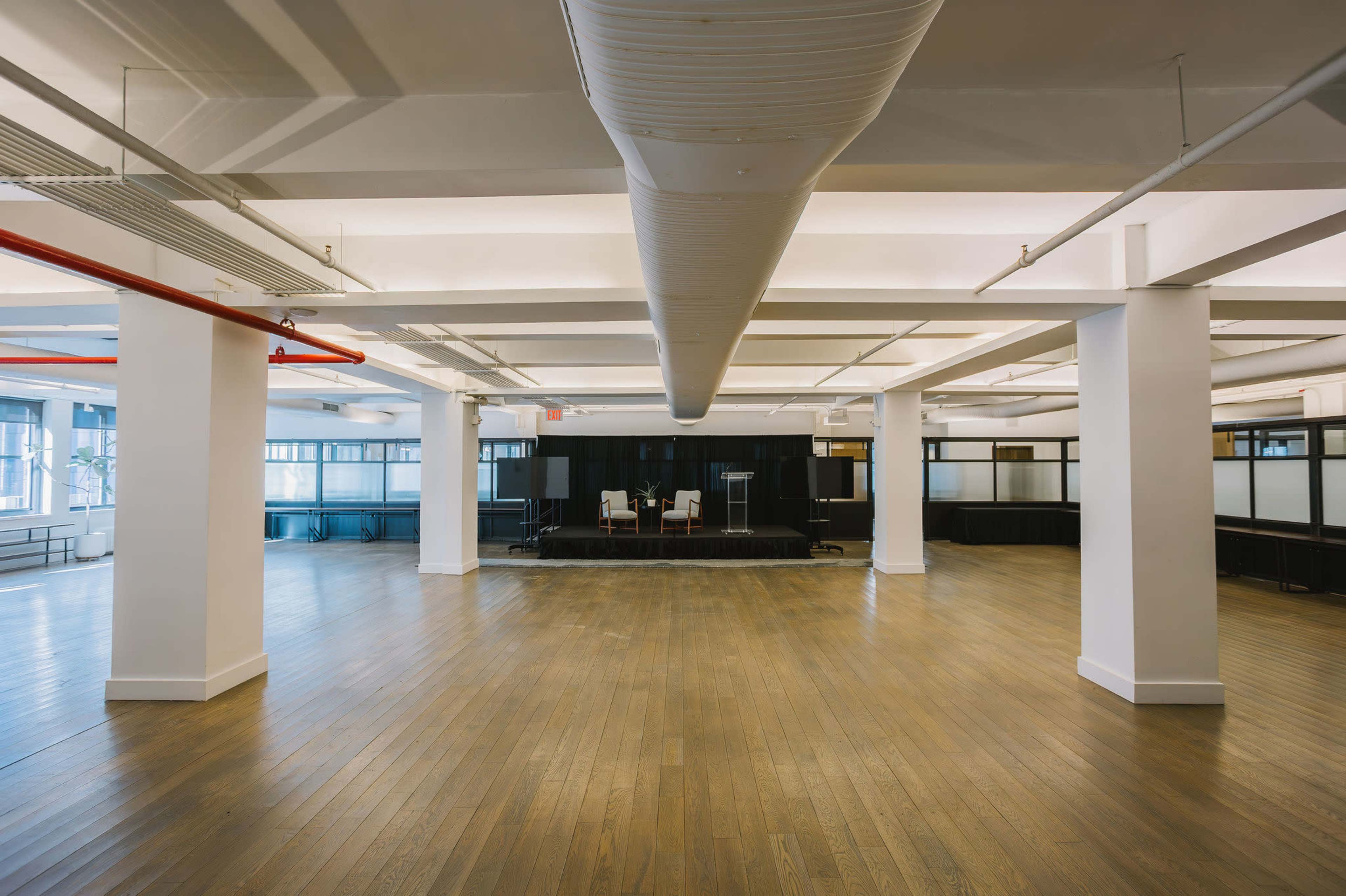 The image shows a spacious, empty office area with wooden flooring, large windows, and a small stage setup at the far end.