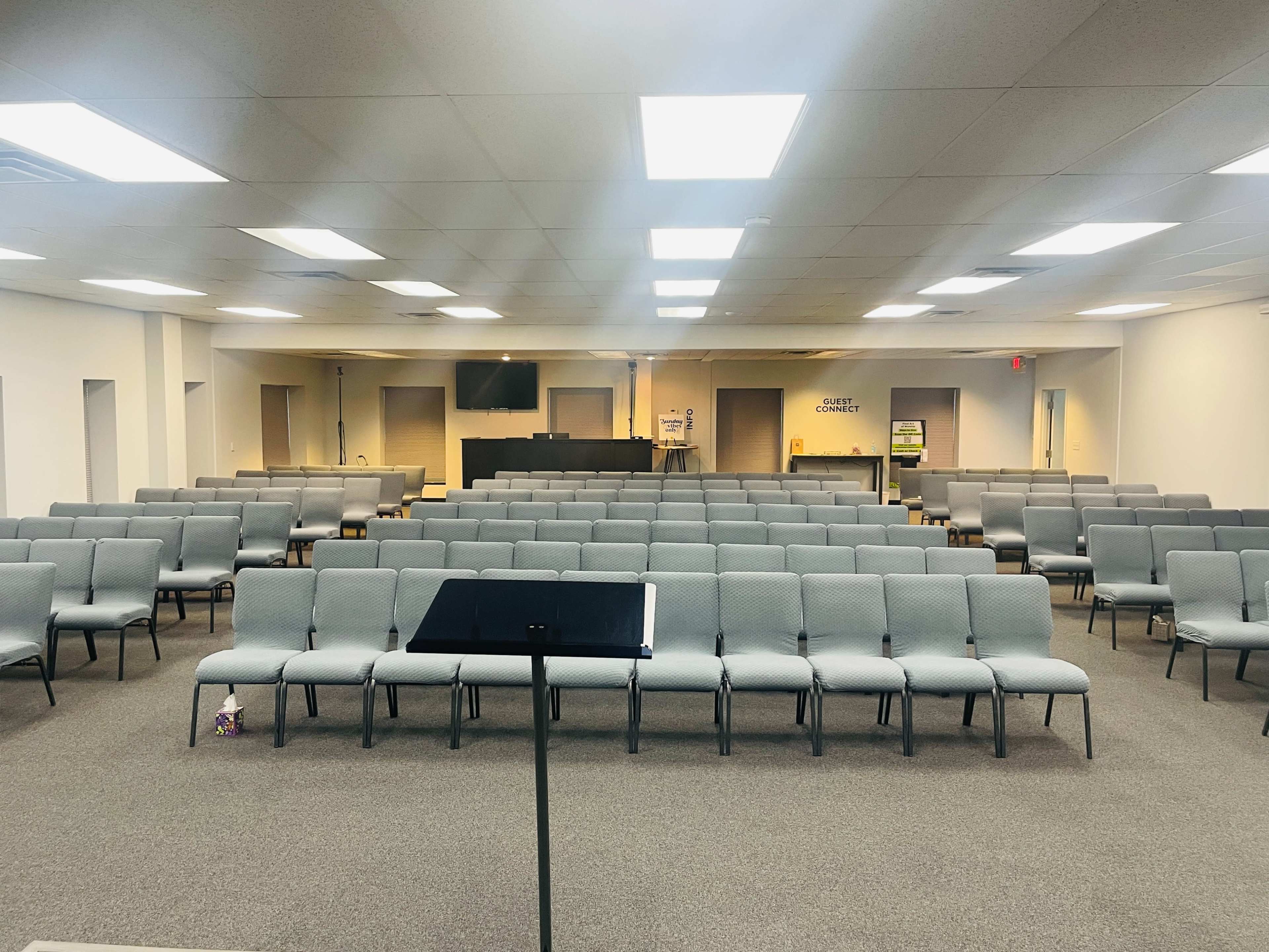 A spacious interior of a hall filled with gray chairs arranged in rows facing a front stage area with a podium and a television.
