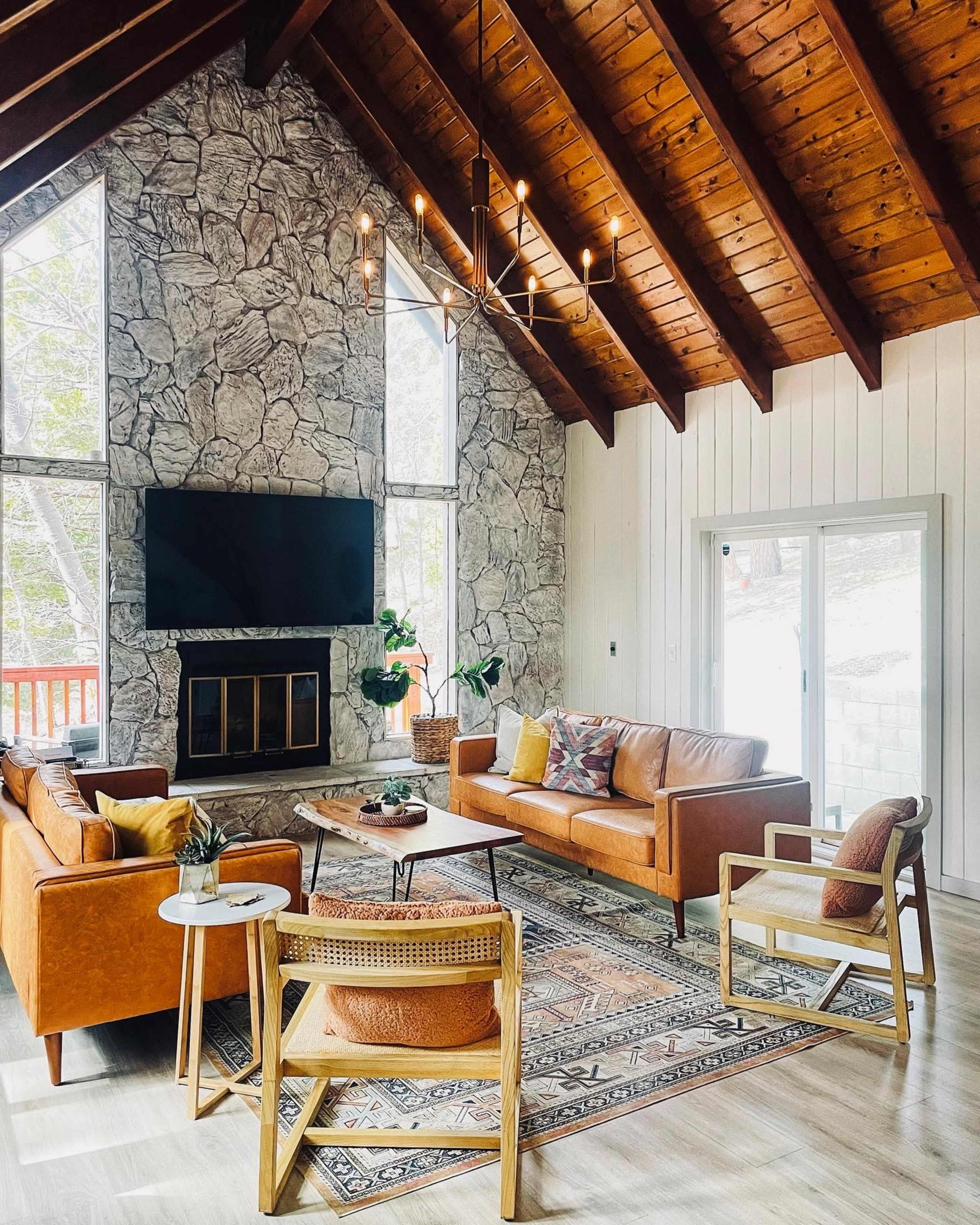 The image shows a modern living room with a stone wall, a large television, leather sofas, a wooden ceiling, and a sliding glass door leading outside.