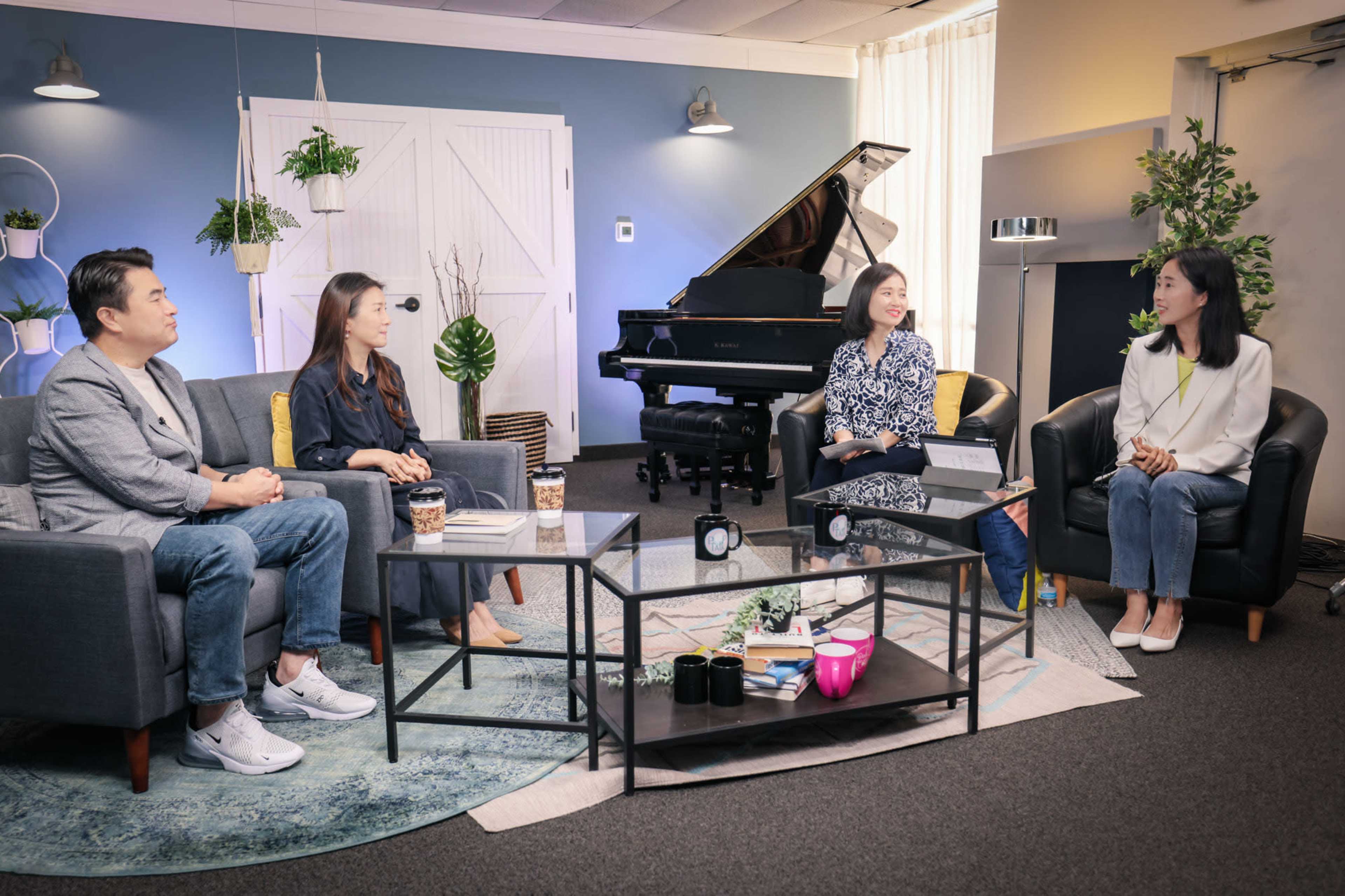 A group of four people sits in a modern interior with a grand piano and plants in the background, engaged in conversation around a coffee table.