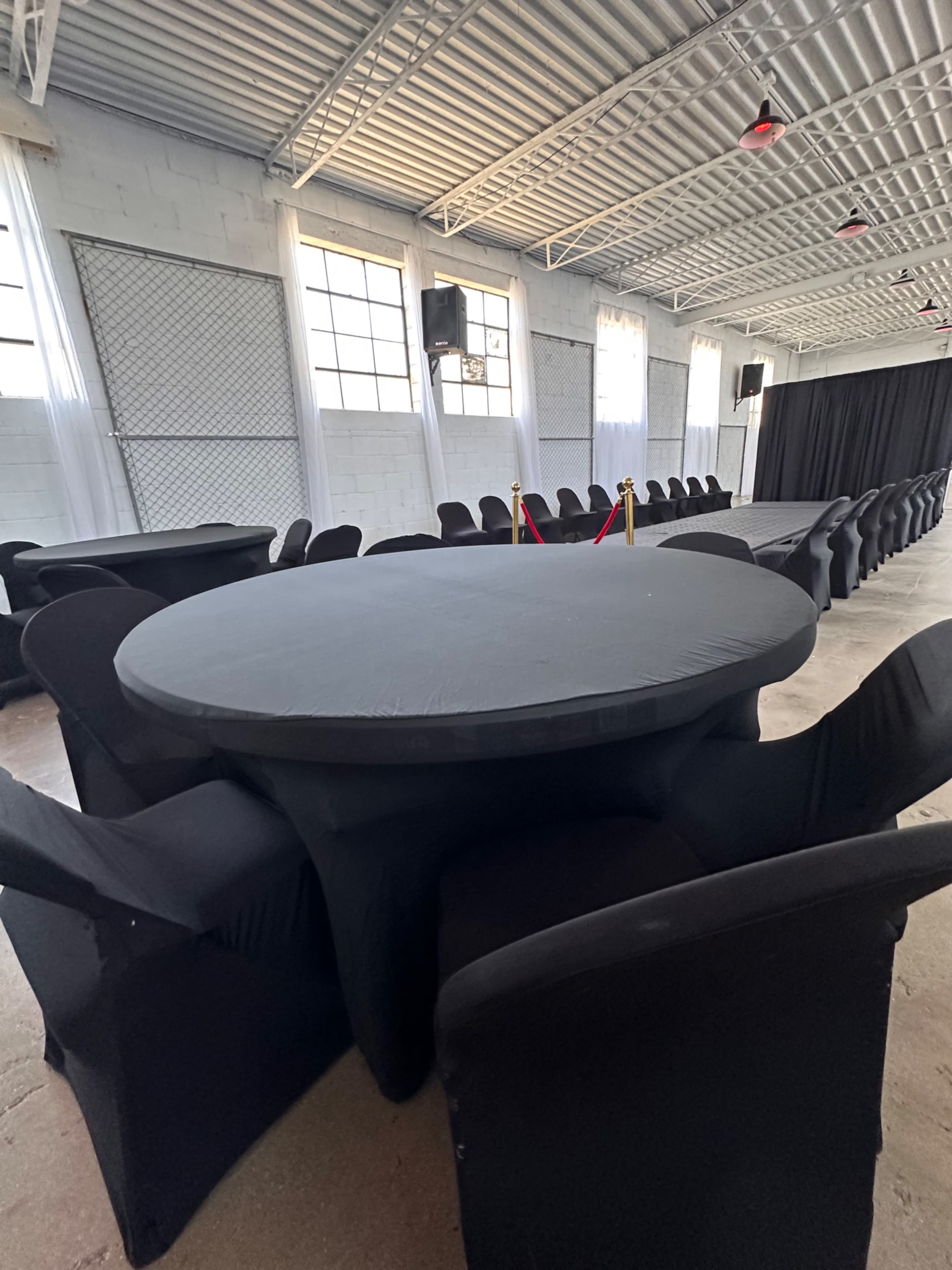 A round table with black covering is surrounded by black chairs in a spacious hall featuring large windows and a stage area.
