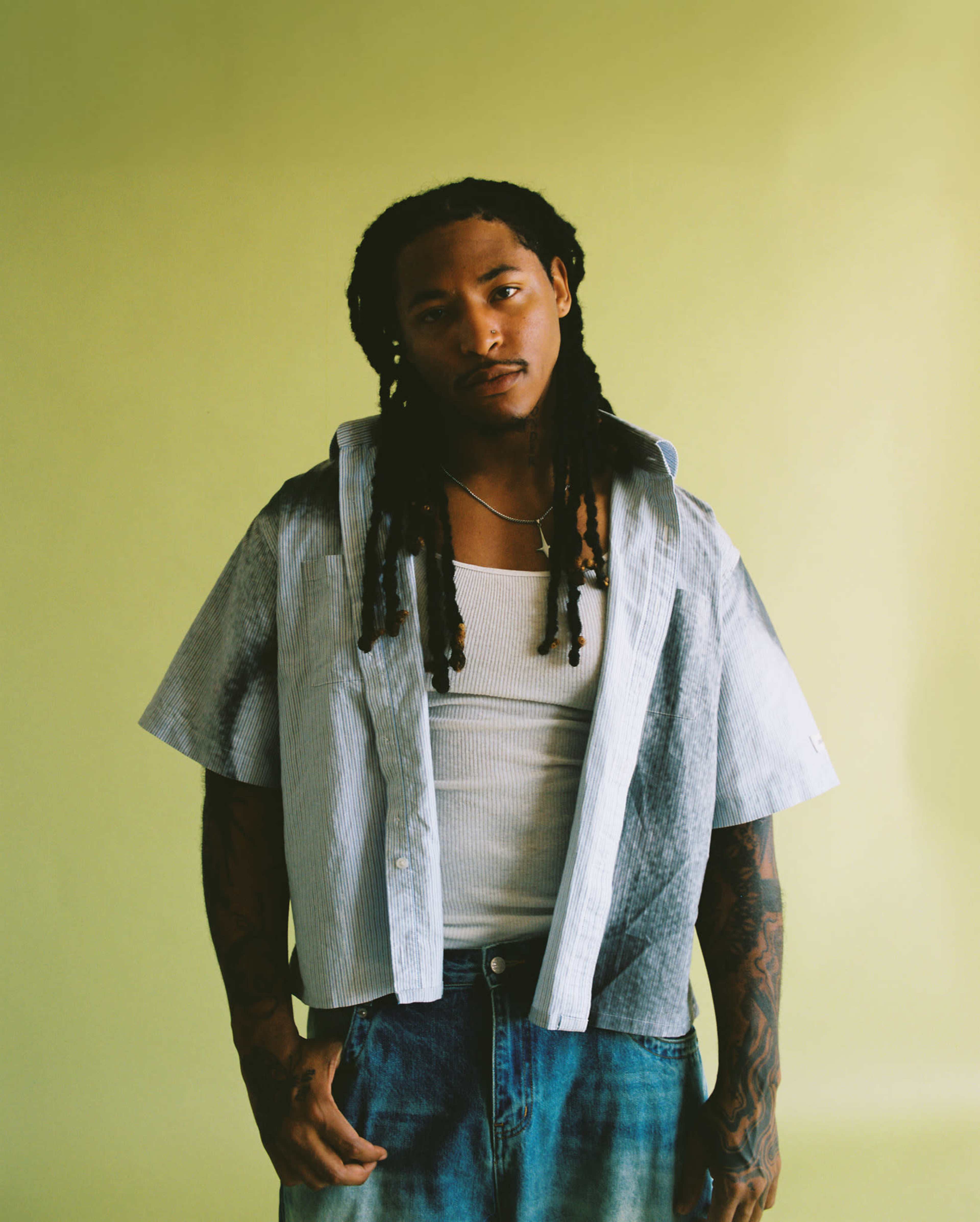 A person with long, braided hair stands against a plain yellow background, wearing a short-sleeved shirt over a white tank top and denim jeans.