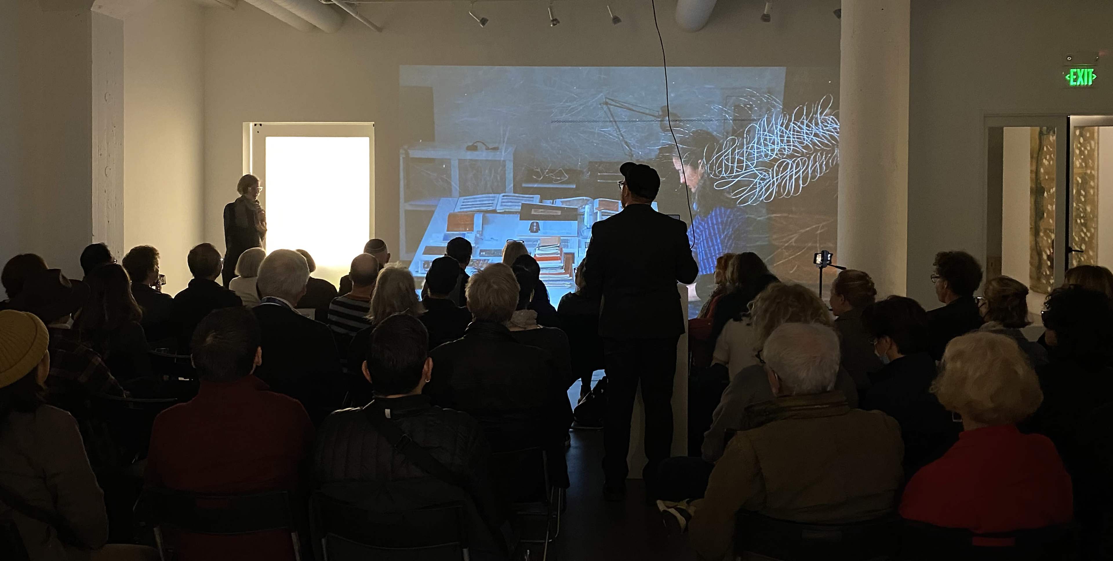 A audience watches a presentation featuring projected visuals in a well-lit gallery space.