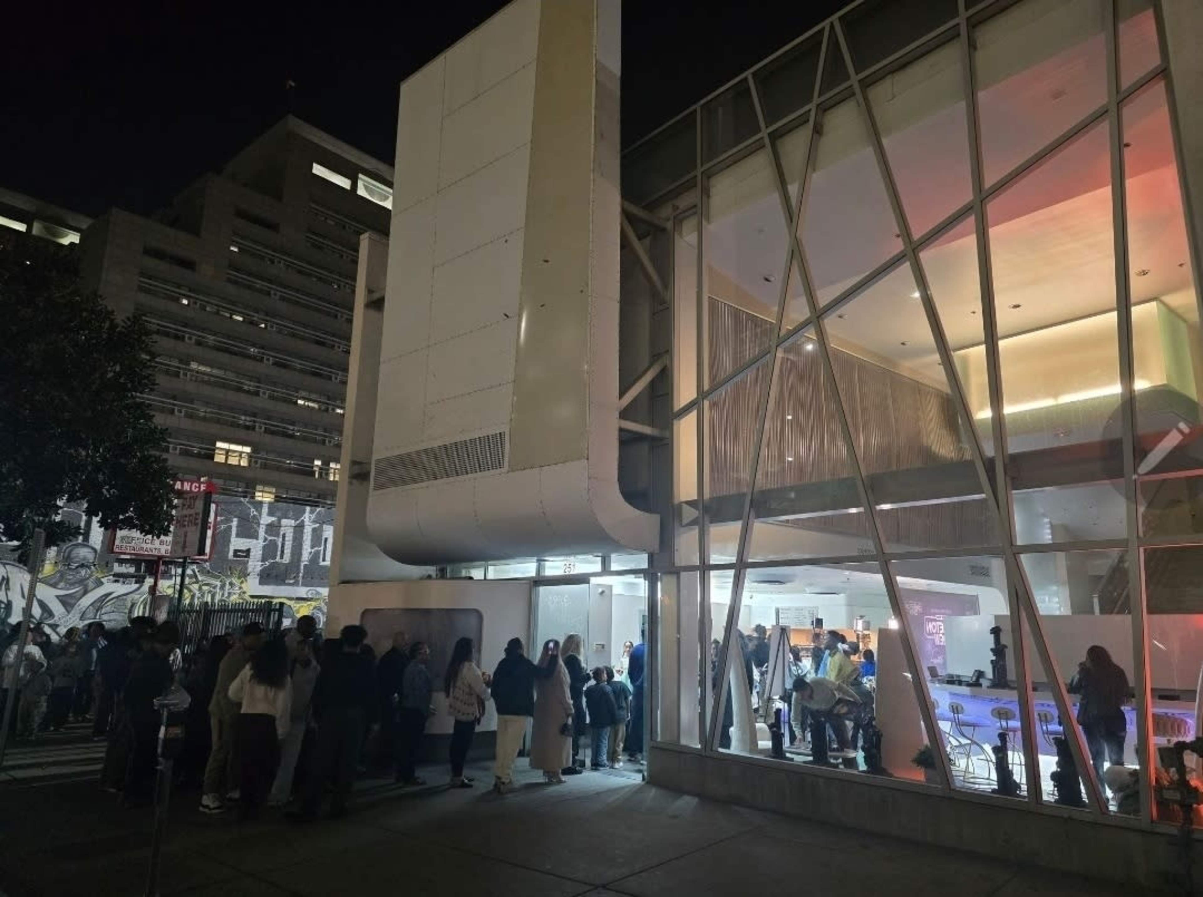 A crowd of people is waiting in line outside a brightly lit modern building at night.