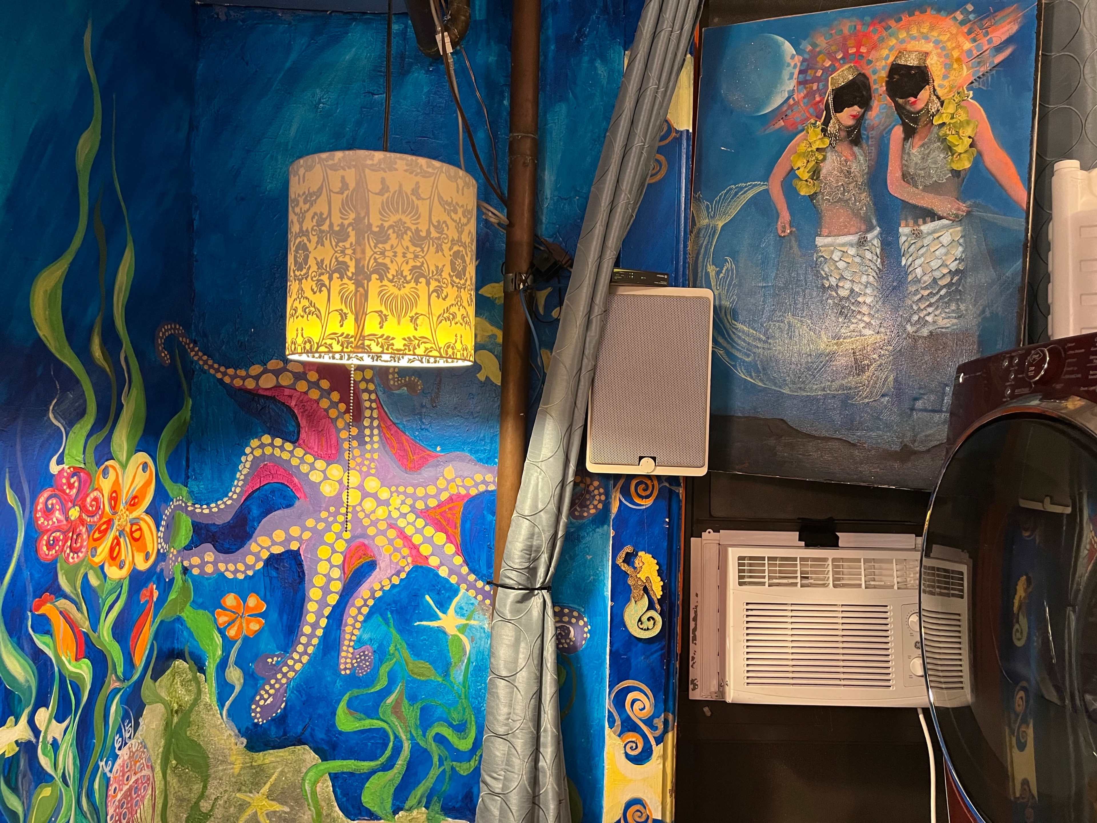 A vibrant mural depicting an octopus and underwater scenes, alongside a painting of two mermaids, all set in a room with a decorative lamp.