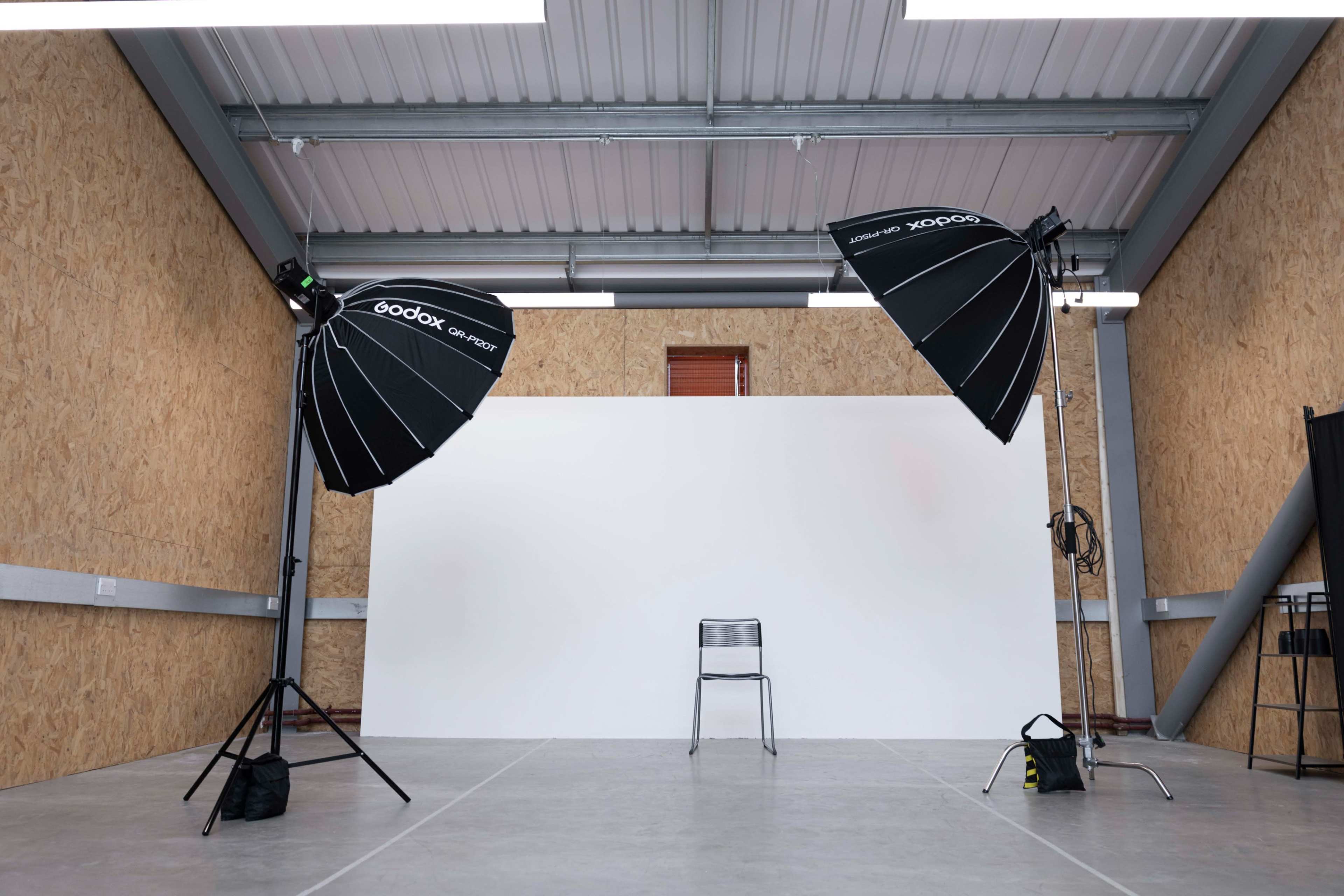 A empty photography studio features a white backdrop and two large softbox lights positioned on either side, with a single chair in the center.