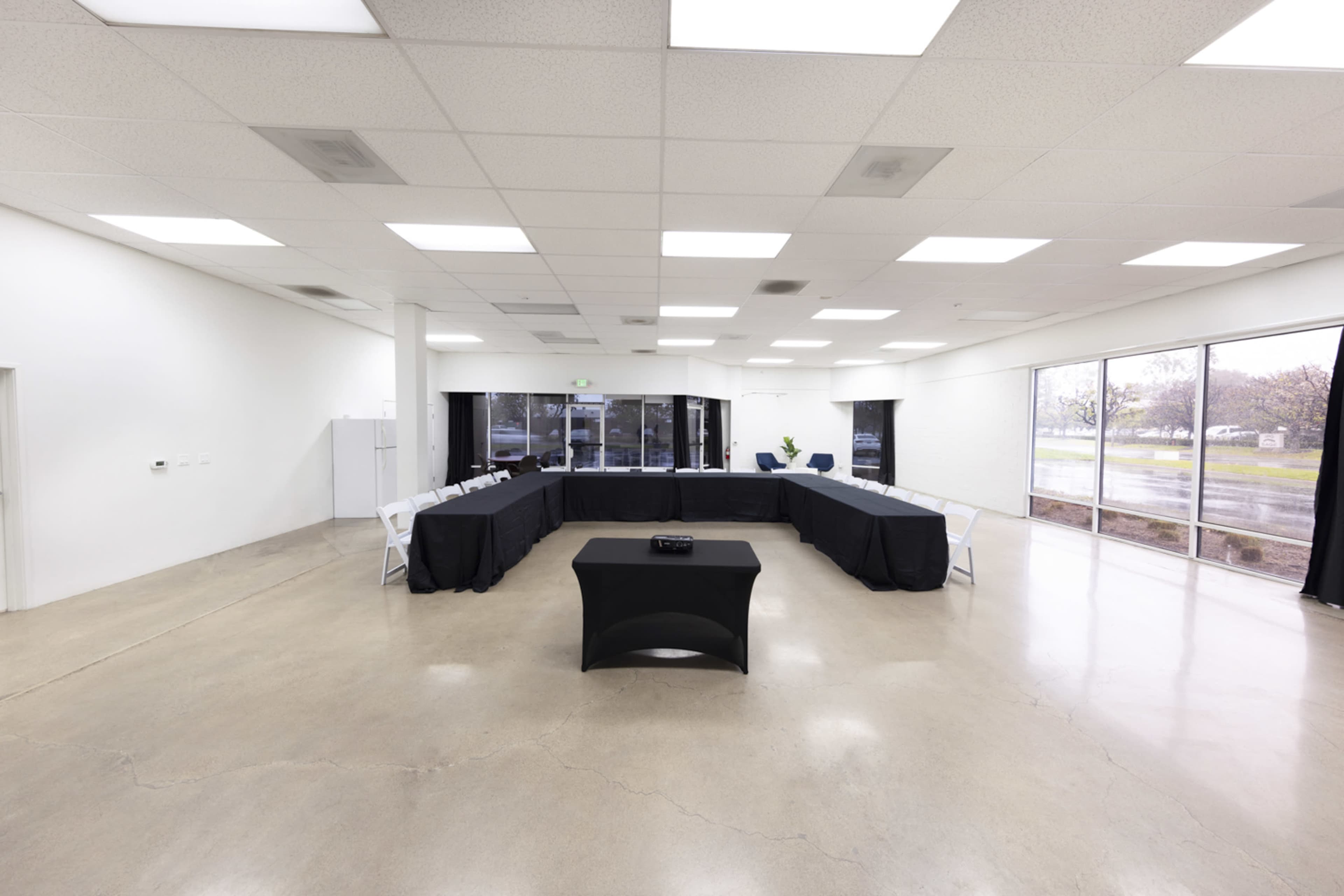 A large, empty conference room features a rectangular table covered with a black cloth, surrounded by chairs, and has large windows letting in natural light.