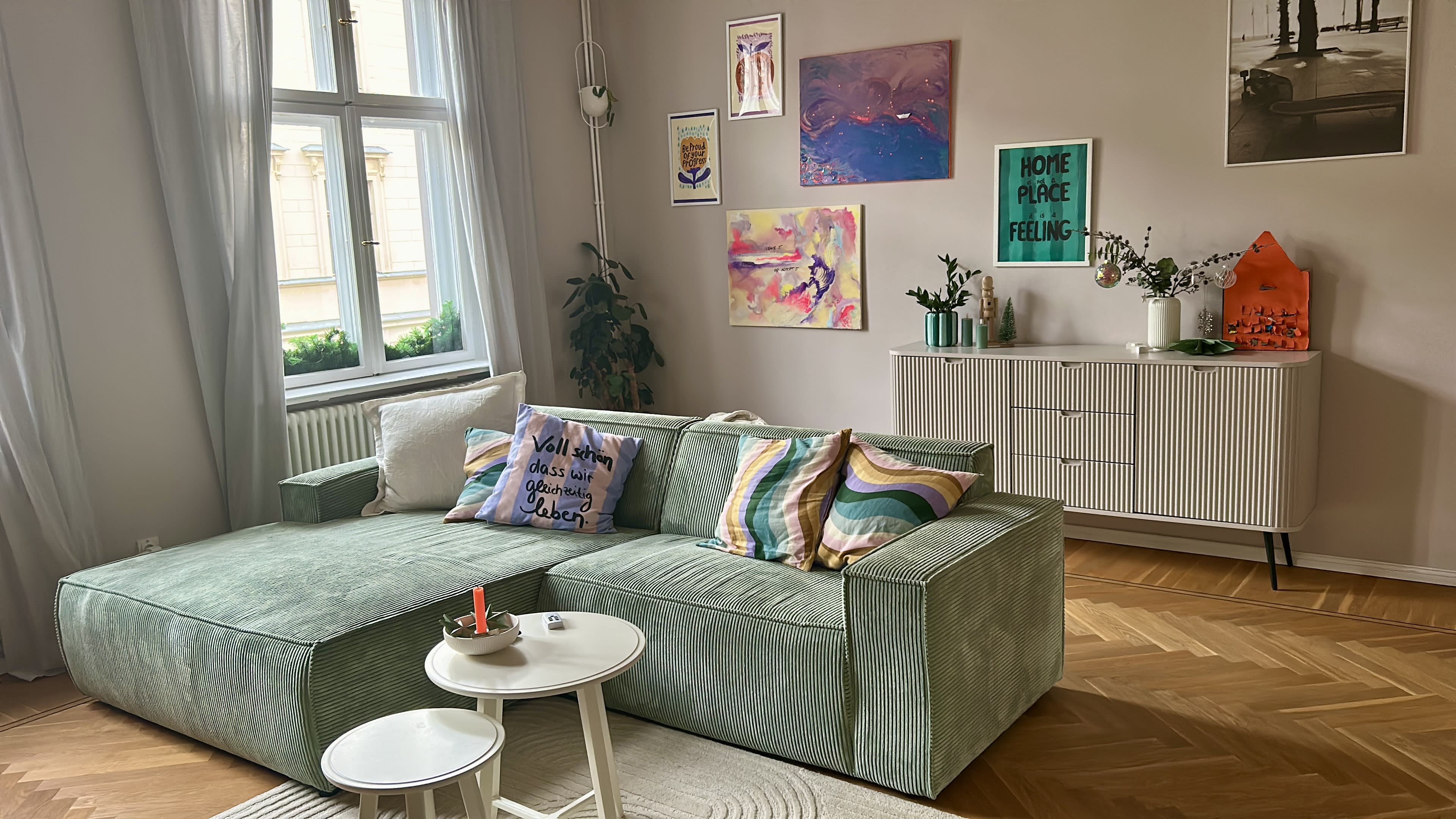 The image shows a bright, modern living room featuring a green sectional sofa, a white side table with a candle, and various pieces of artwork on the walls.