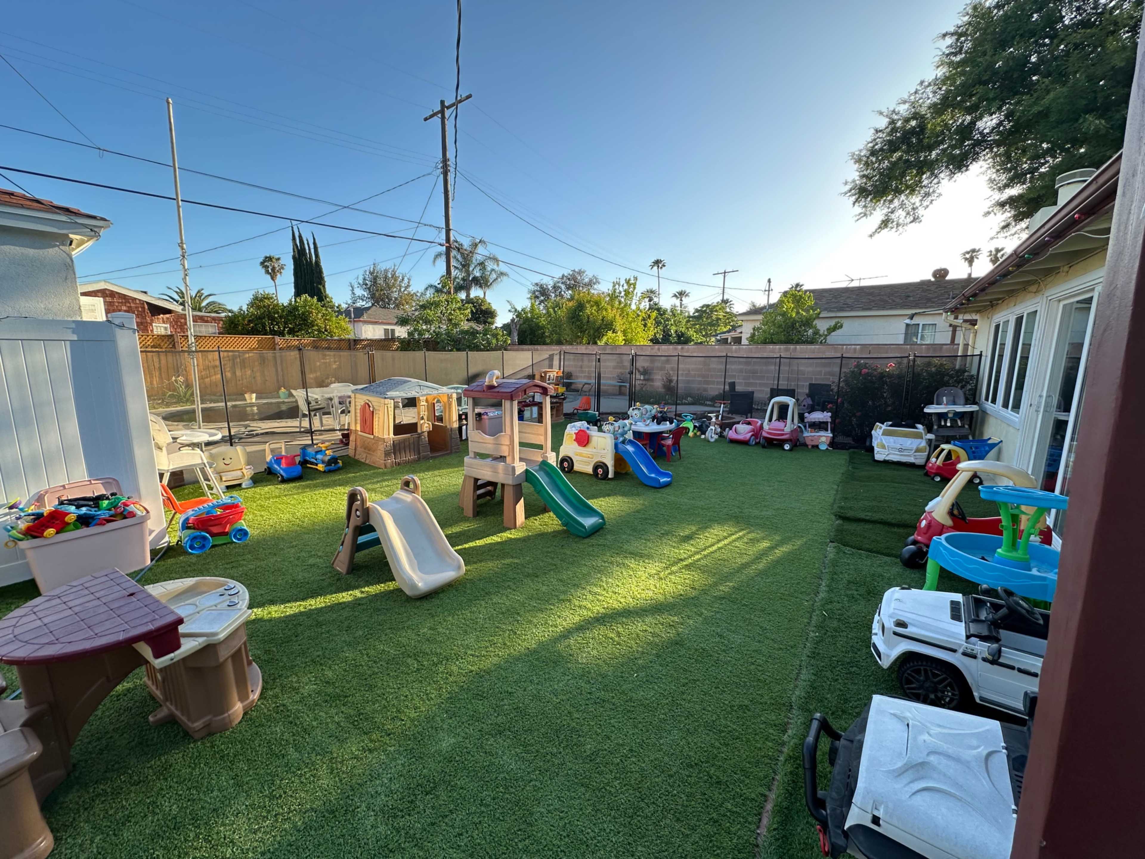 The image shows a playful outdoor space with various children’s toys and play structures on a green turf lawn.