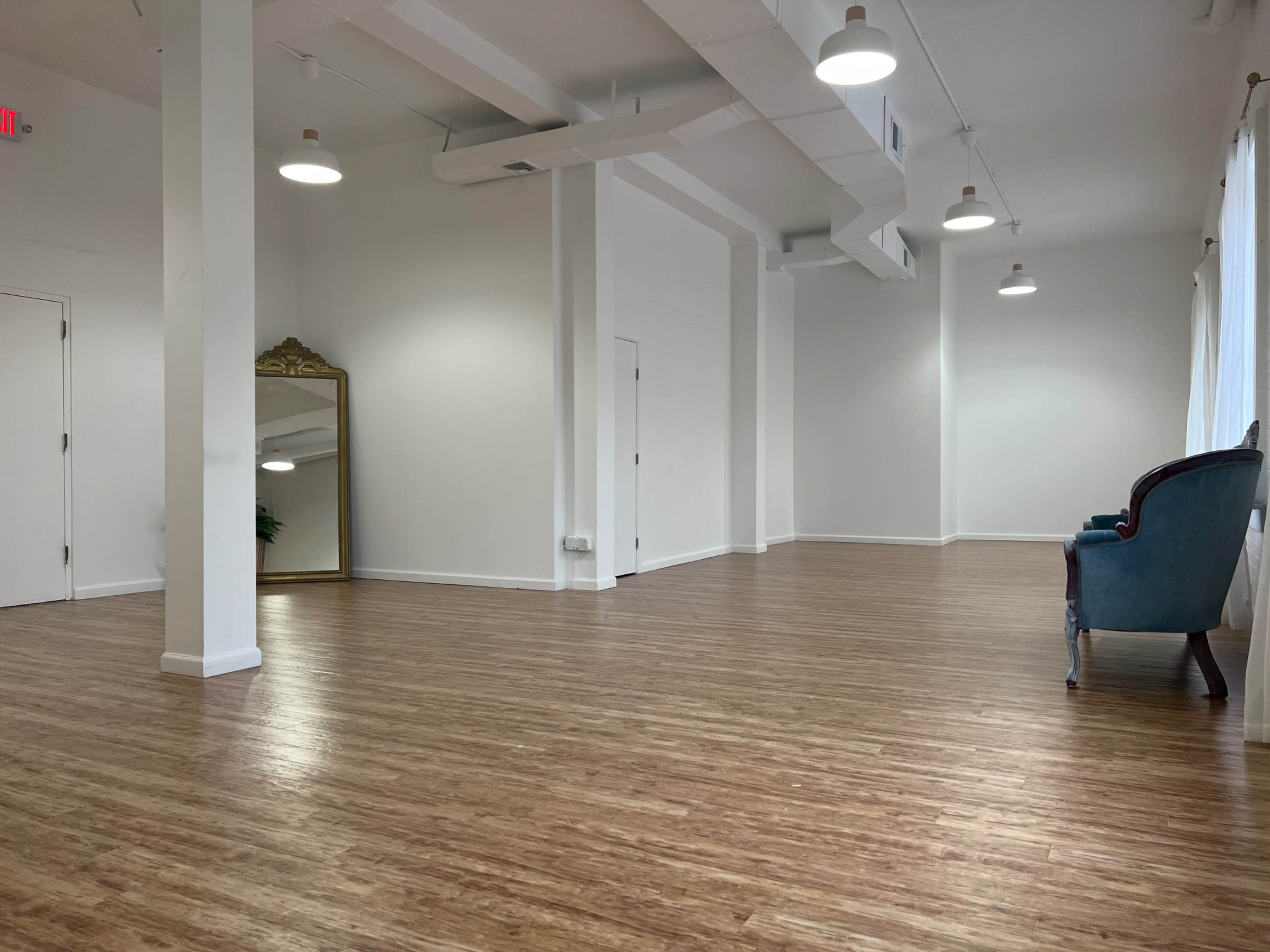 The image shows a spacious, empty room with wooden flooring, white walls, and a large mirror on one side, complemented by a teal armchair.