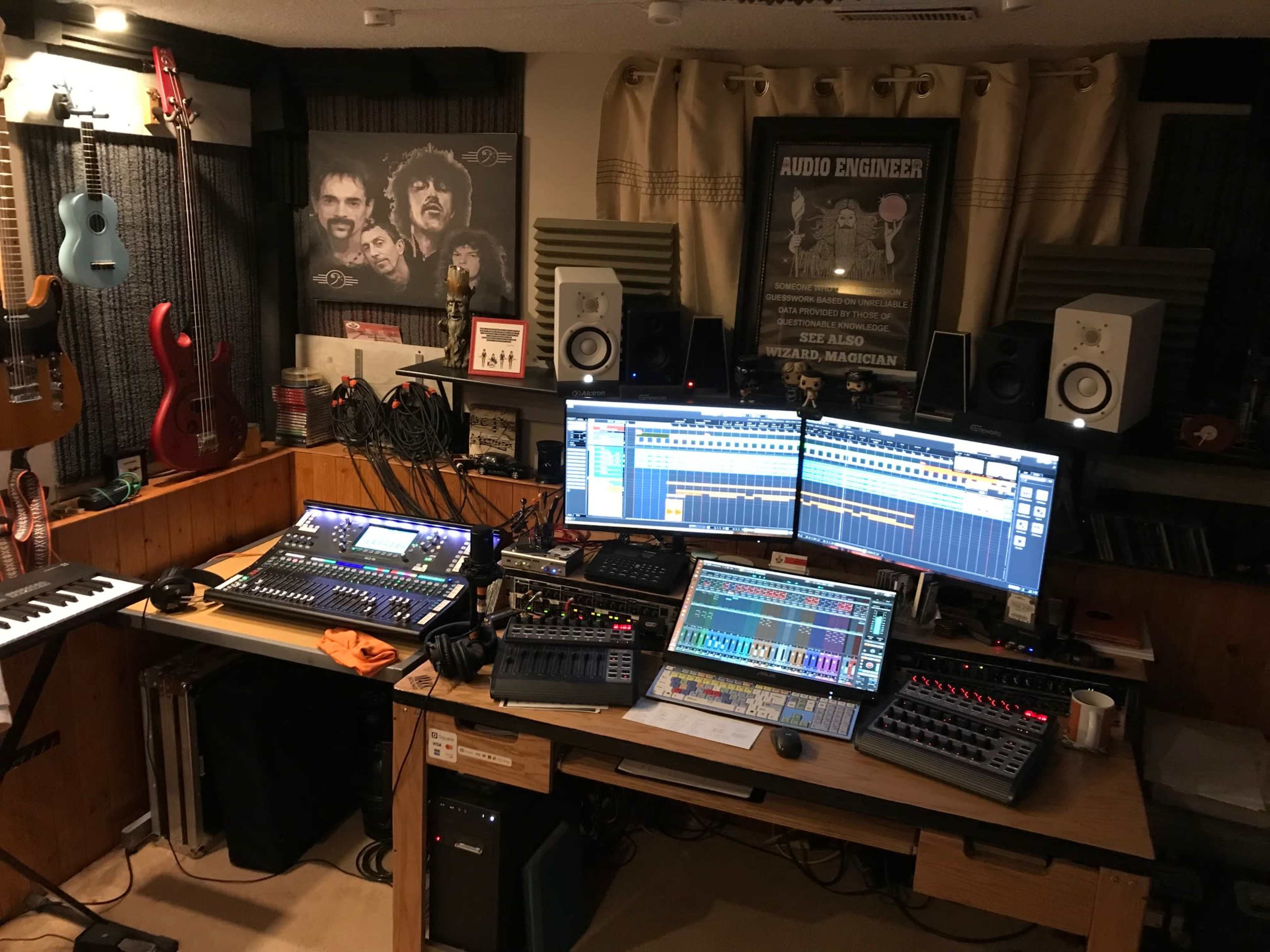 The image shows a music recording studio with multiple audio equipment, instruments, and two large computer monitors displaying sound editing software.