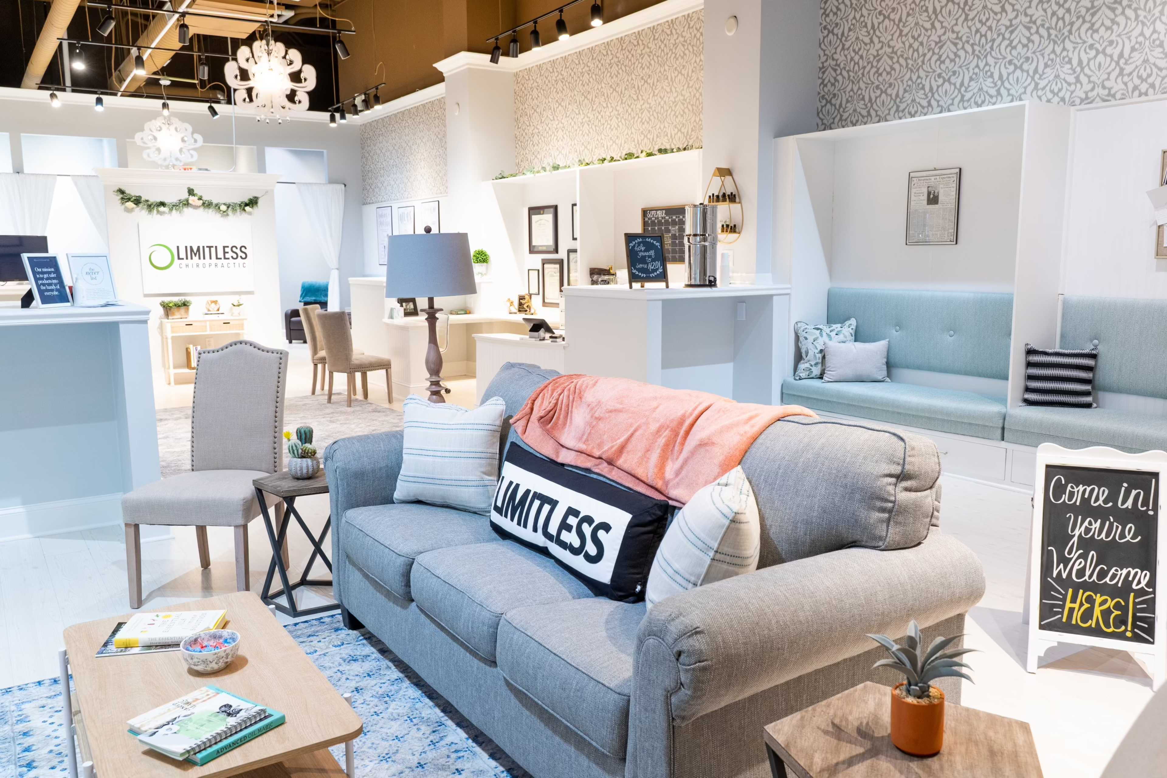 A modern showroom featuring a grey sofa with decorative pillows, a wooden coffee table with books, and a welcoming sign, set against light walls and stylish decor.