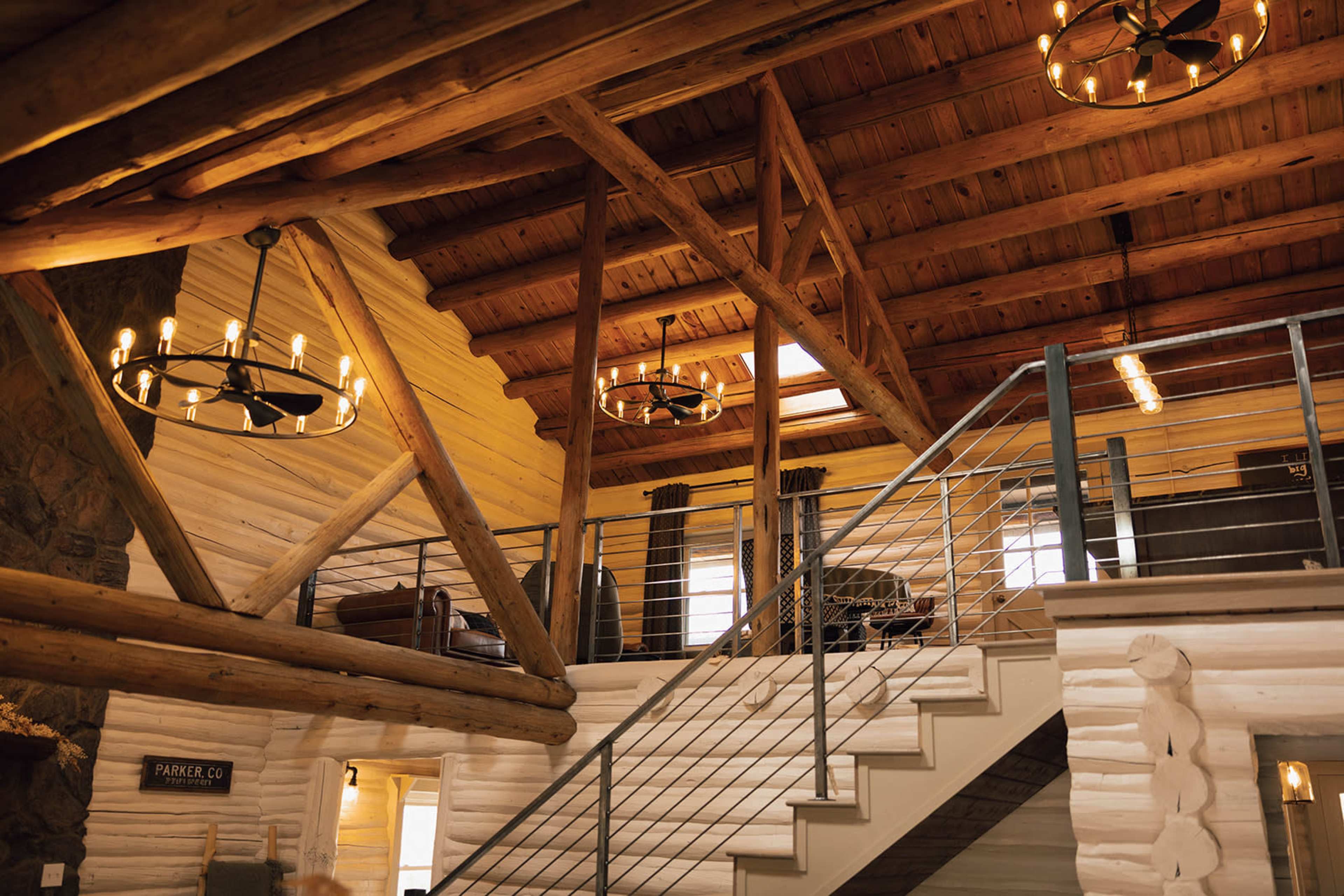 The photo showcases the interior of a rustic log cabin featuring wooden beams, a staircase, and industrial-style lighting fixtures.