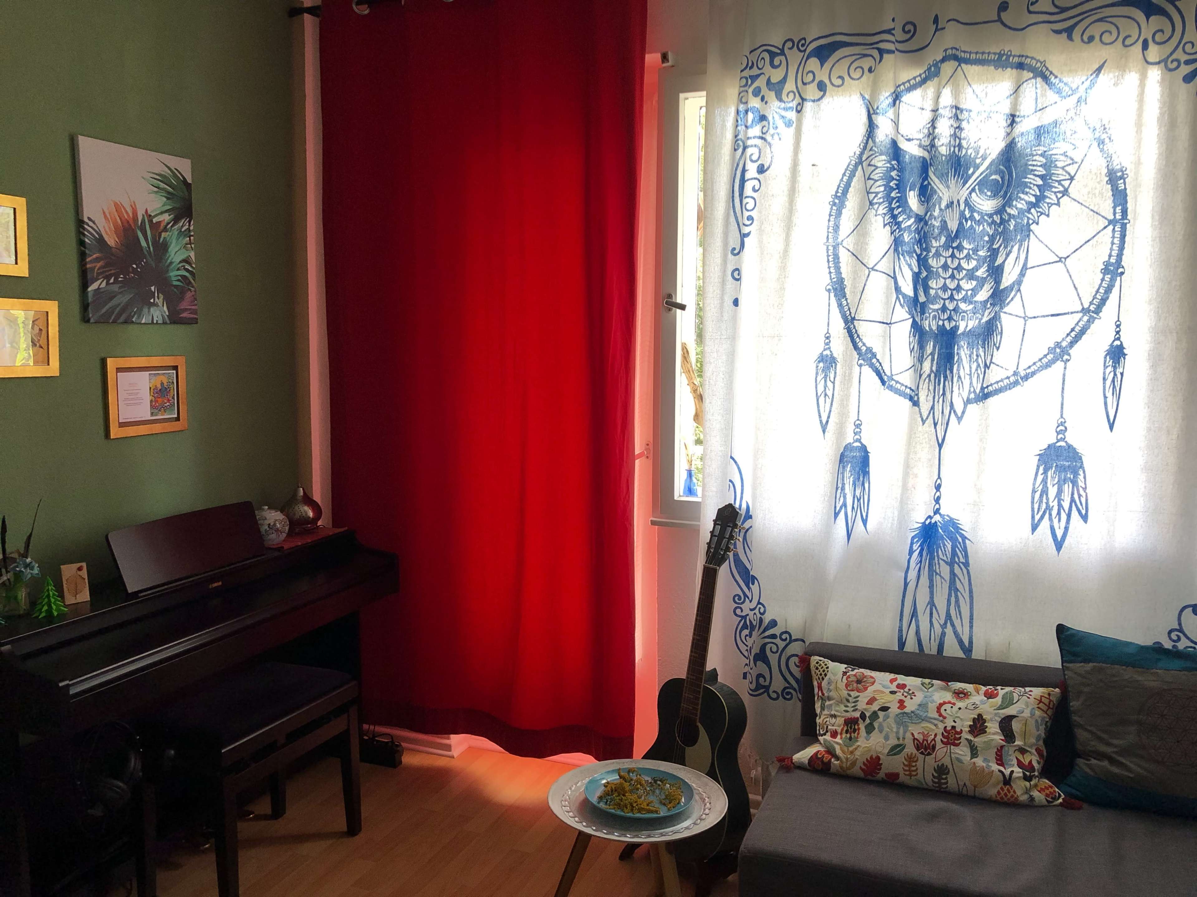 A cozy room features a dark piano, a red curtain, a decorative owl tapestry on the window, and a plate of food on a side table next to a sofa with patterned cushions and an acoustic guitar.