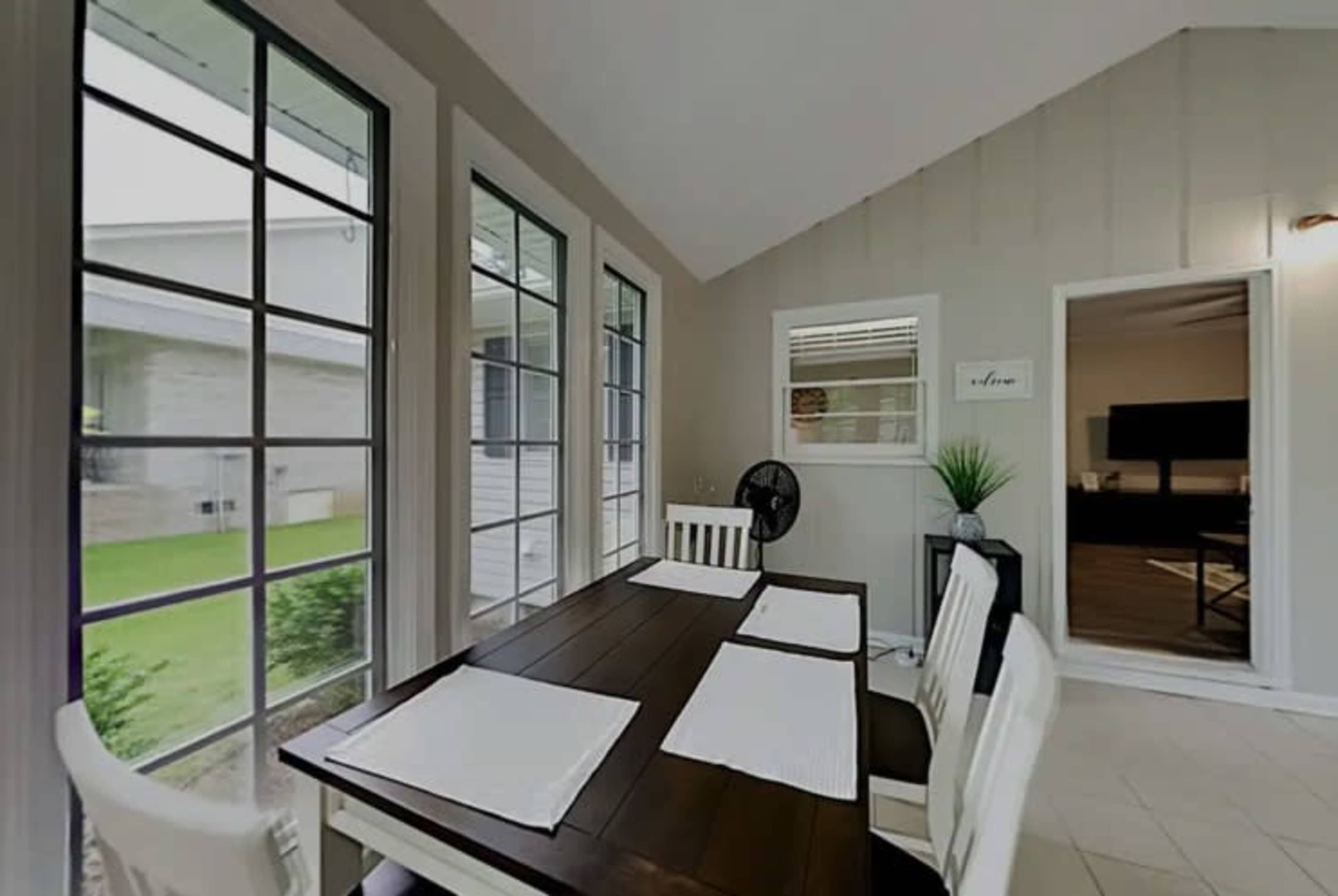 The image shows a dining area with a table and chairs, surrounded by large windows, leading to an adjoining room.