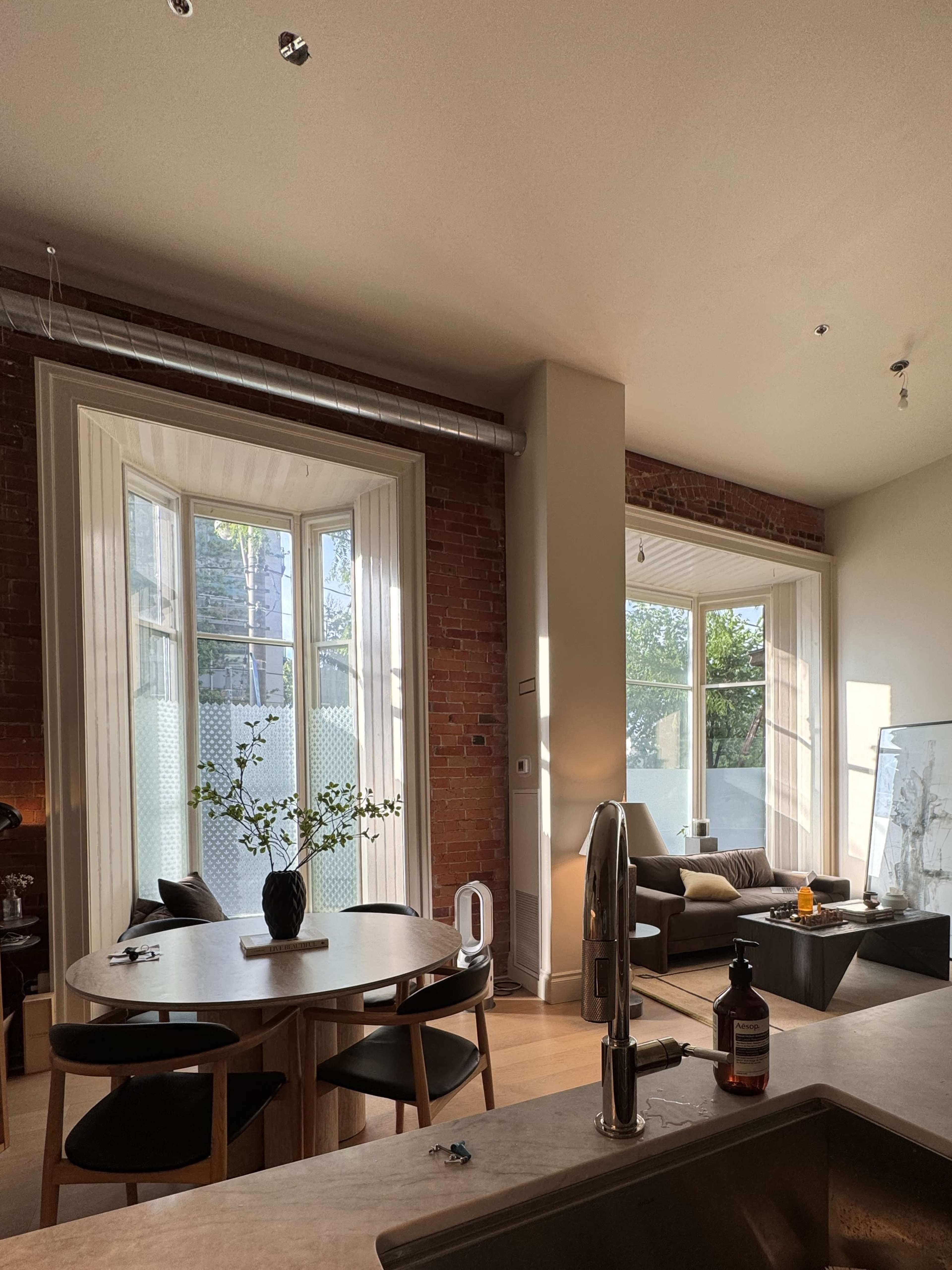 The image shows a modern living space with large windows, a dining area featuring a round table and chairs, and a comfortable seating area with a couch.