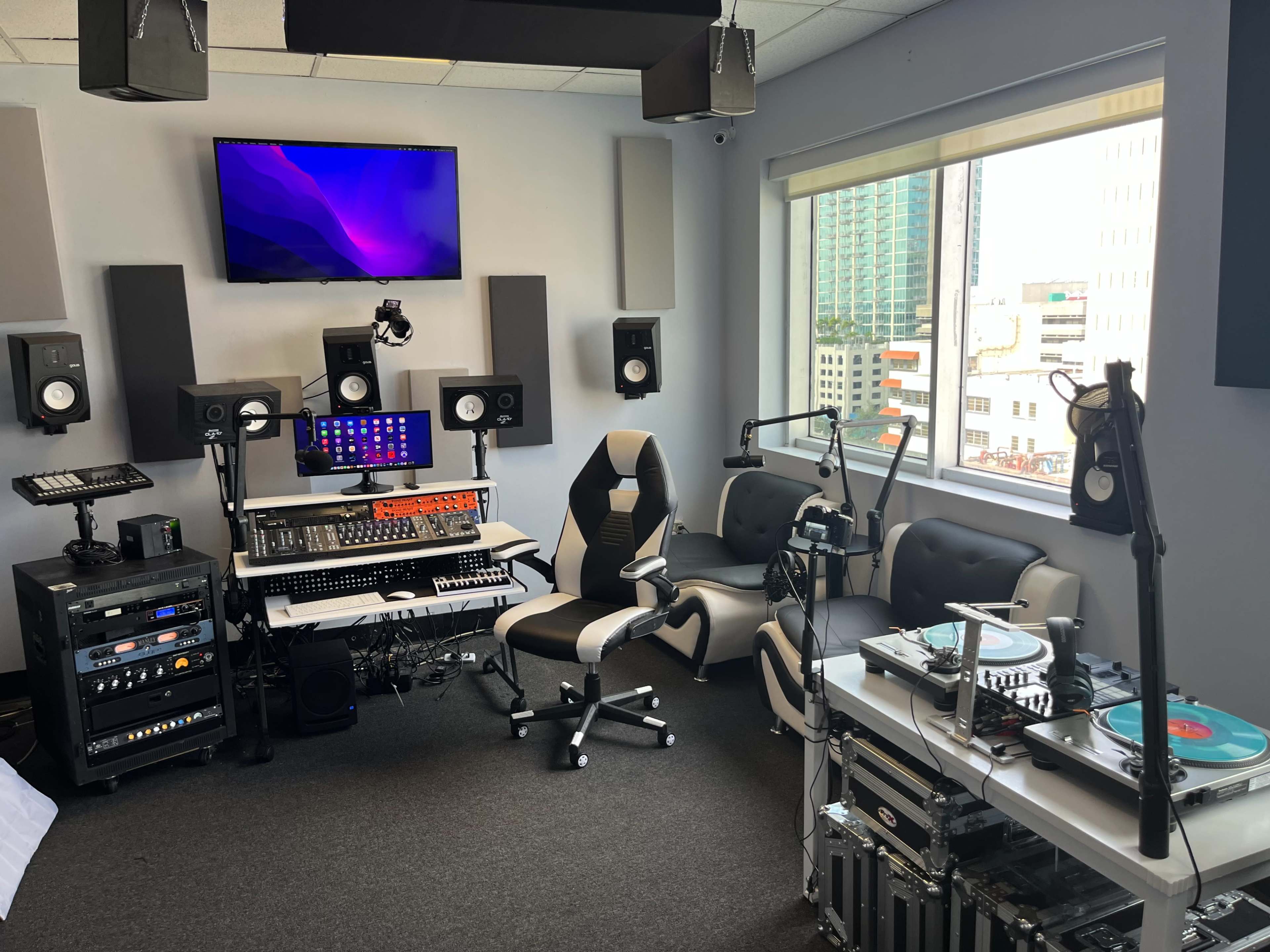 A modern music studio equipped with audio equipment, a computer setup, and seating near a large window overlooking a cityscape.