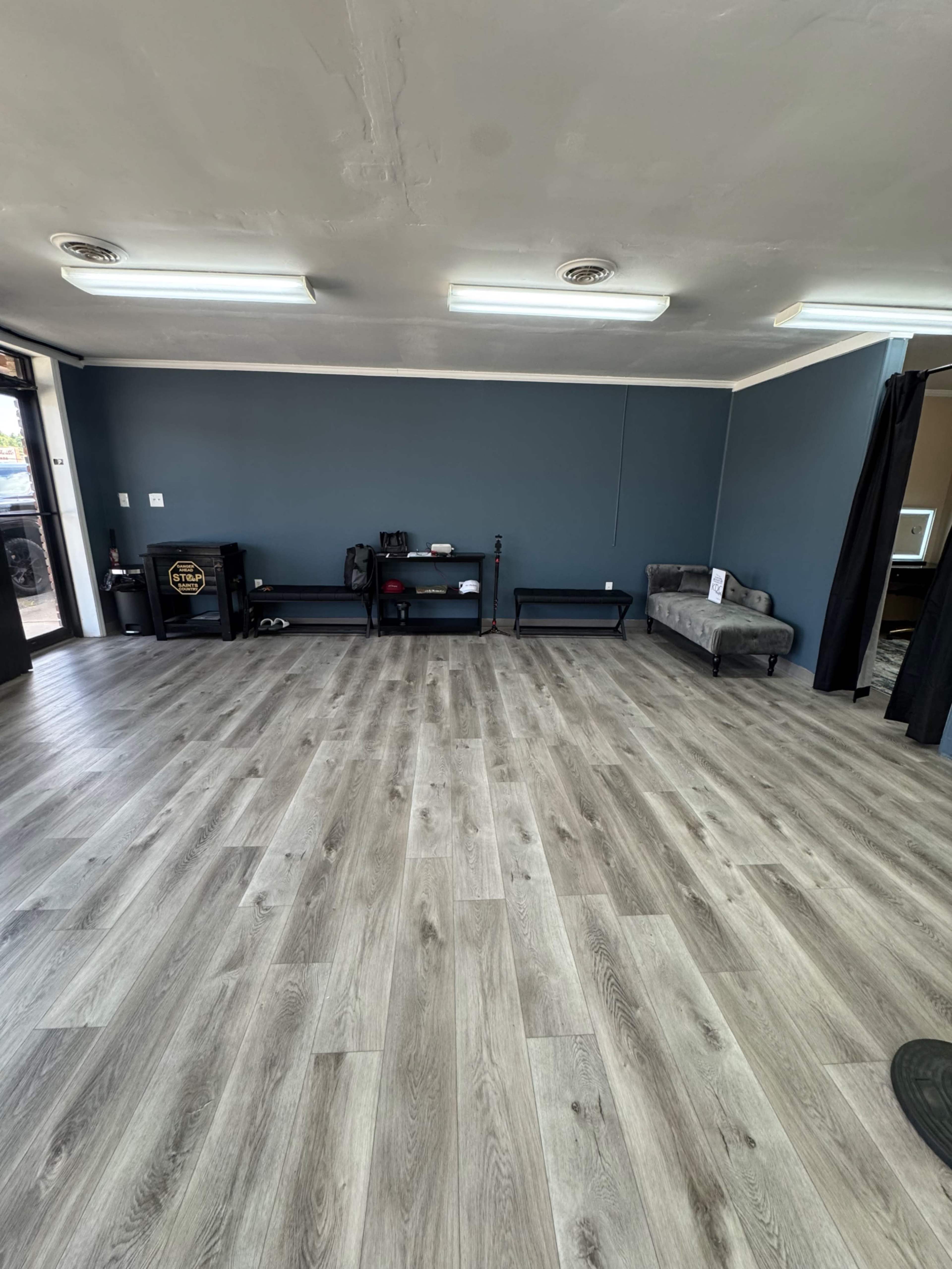 The image shows an empty room with grey wooden flooring, a blue accent wall, a small couch, and minimalist shelving.