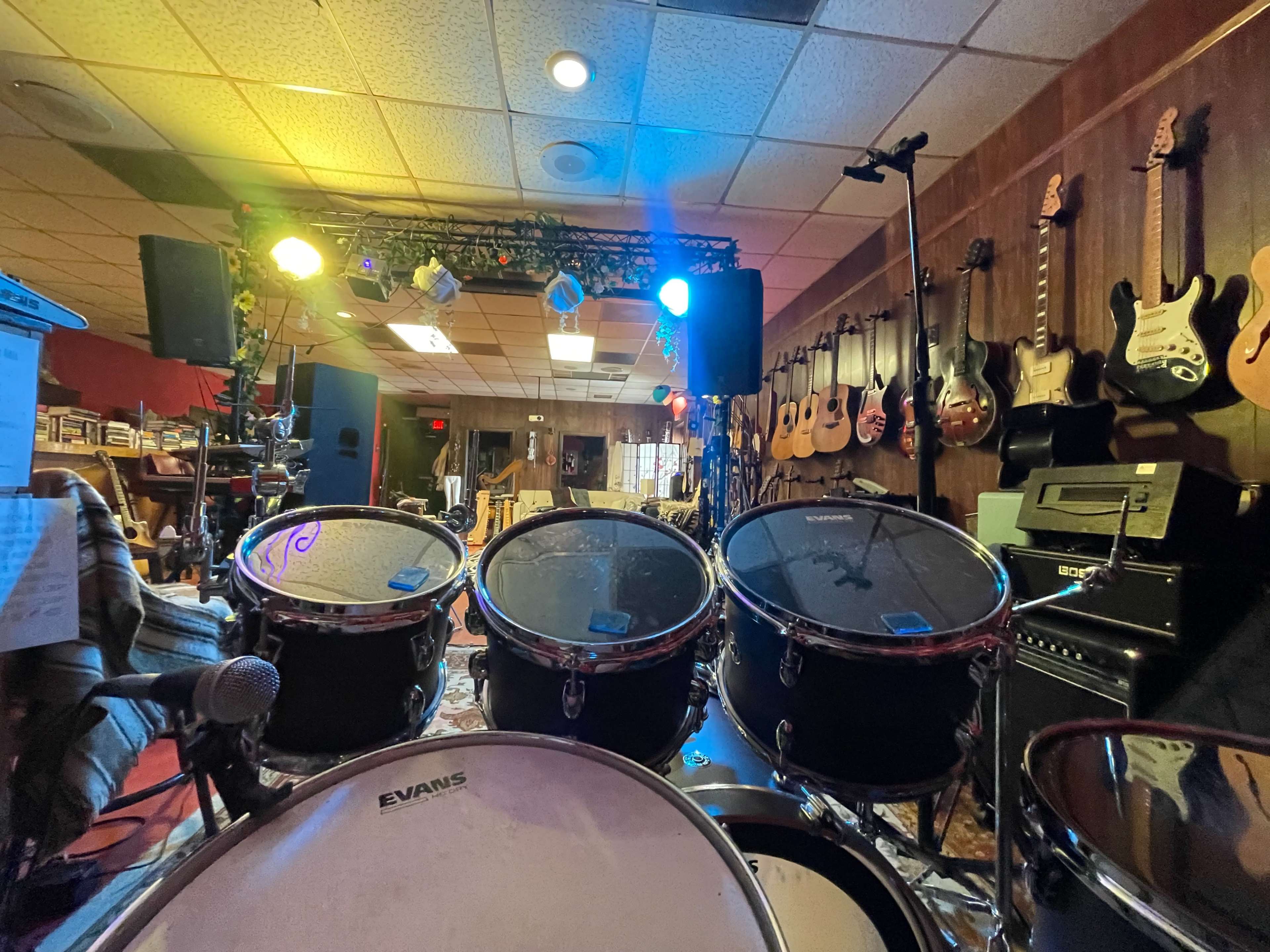 A view from behind a drum kit, with various guitars hanging on the wall and stage lights illuminating the space.