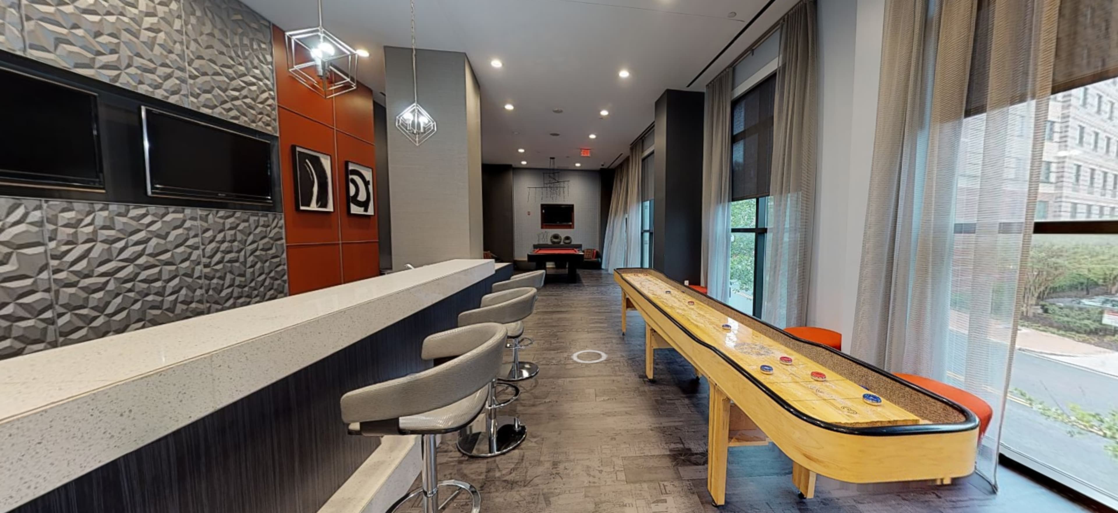 The image shows a modern lounge area featuring a long countertop with bar stools on one side and a shuffleboard table on the other, surrounded by large windows.