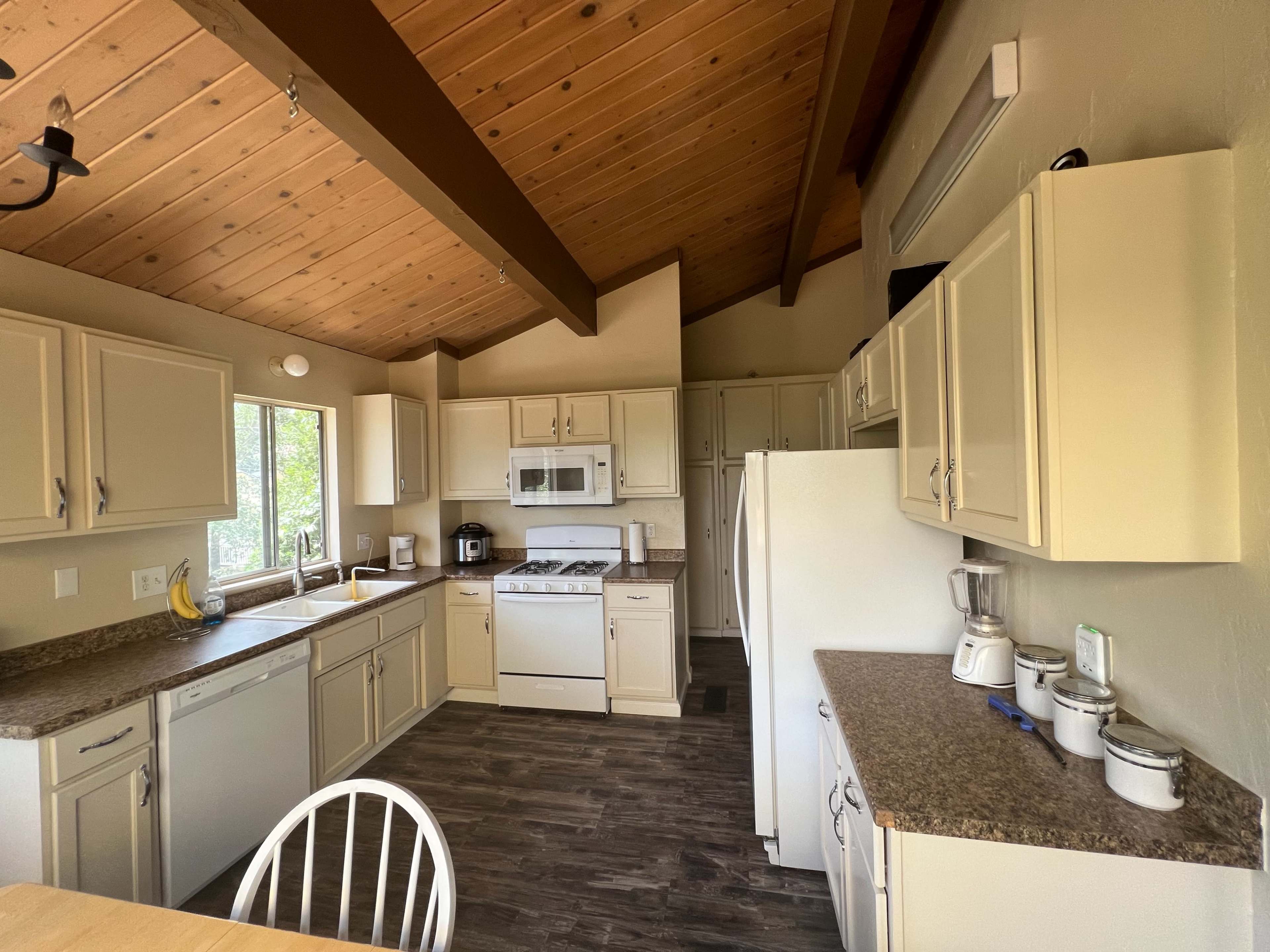 A modern kitchen features light-colored cabinetry, a stove, refrigerator, and countertop appliances, with wooden beams and a window providing natural light.