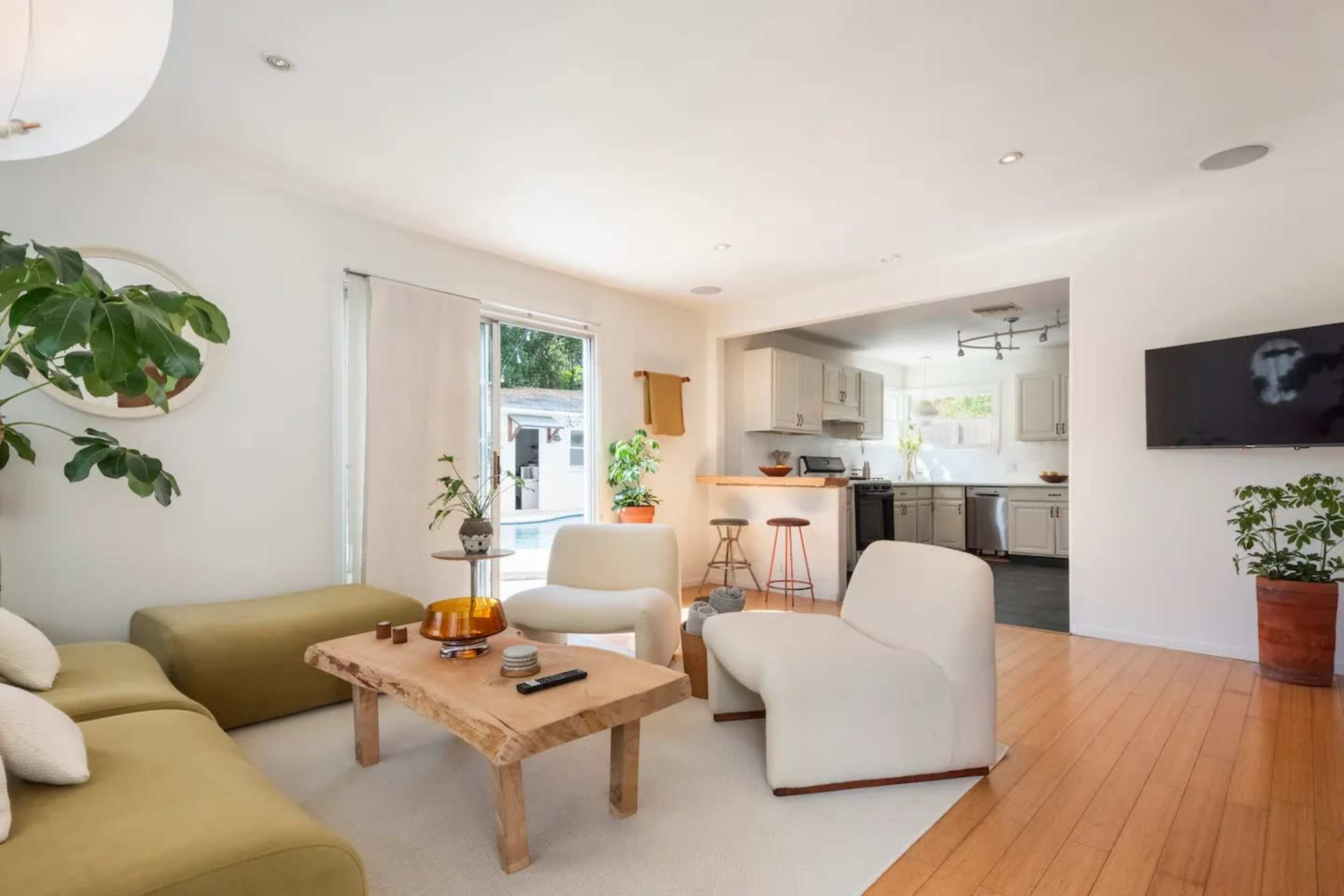 A modern living room with a green couch, white armchairs, a wooden coffee table, and an open kitchen area visible in the background.
