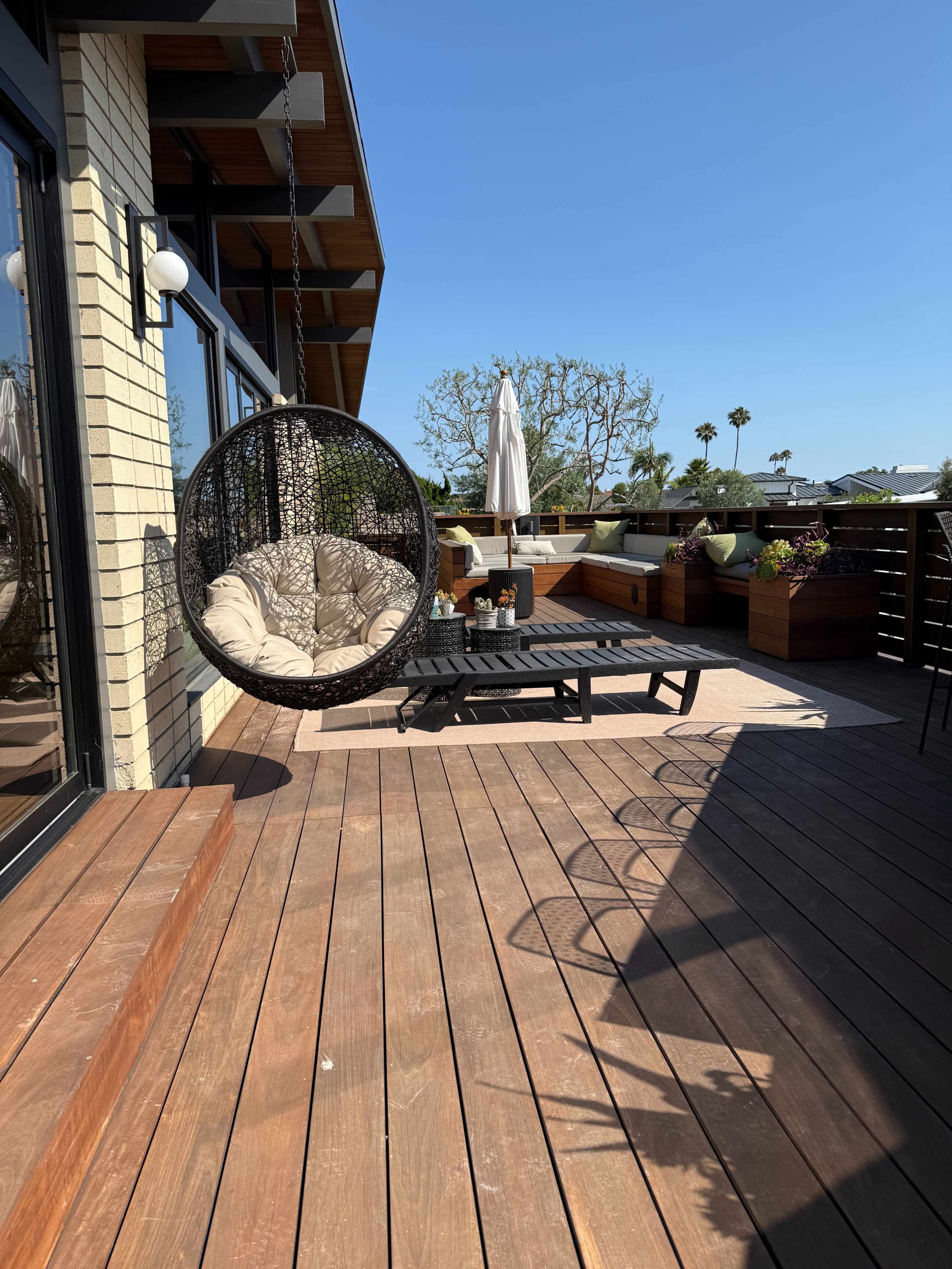 A hanging chair is positioned on a wooden deck, accompanied by a lounge chair and potted plants, against a backdrop of clear blue skies.